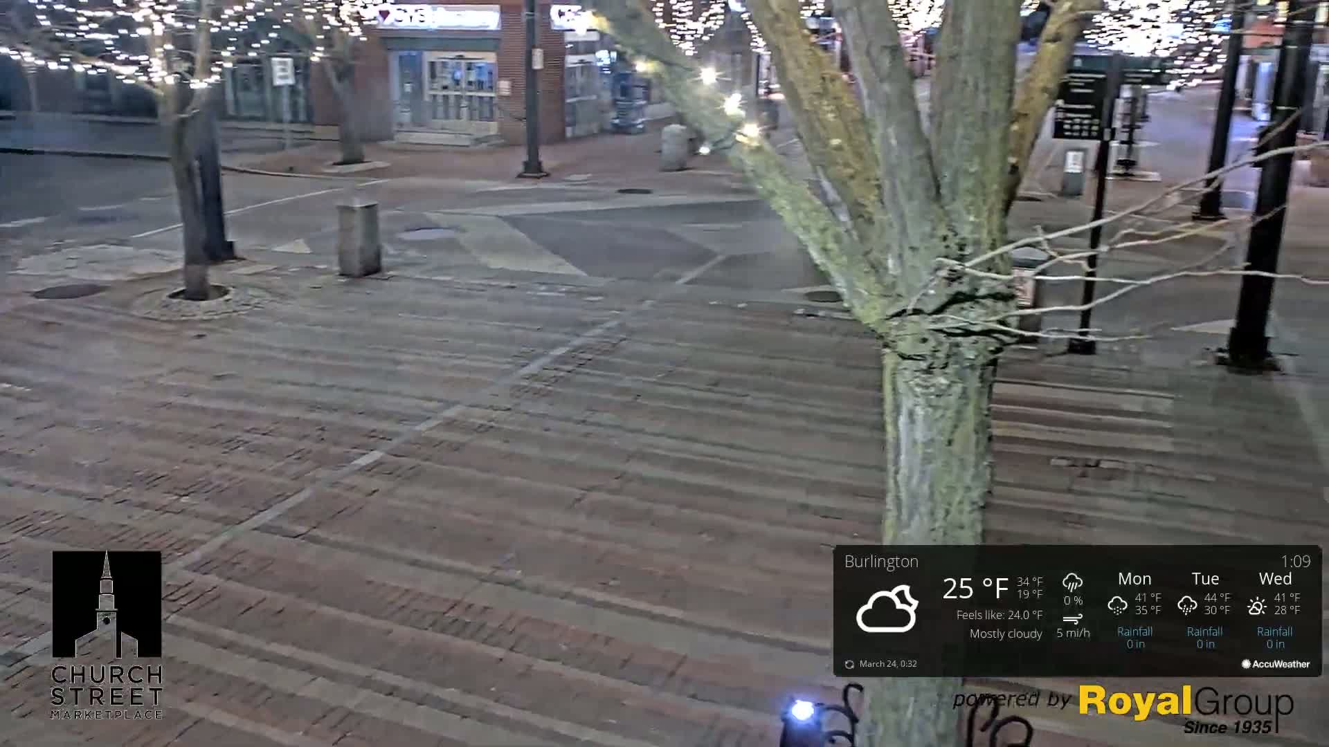 A mostly cloudy night scene shows a brick paved area with a tree in the center, under string lights on nearby branches.
