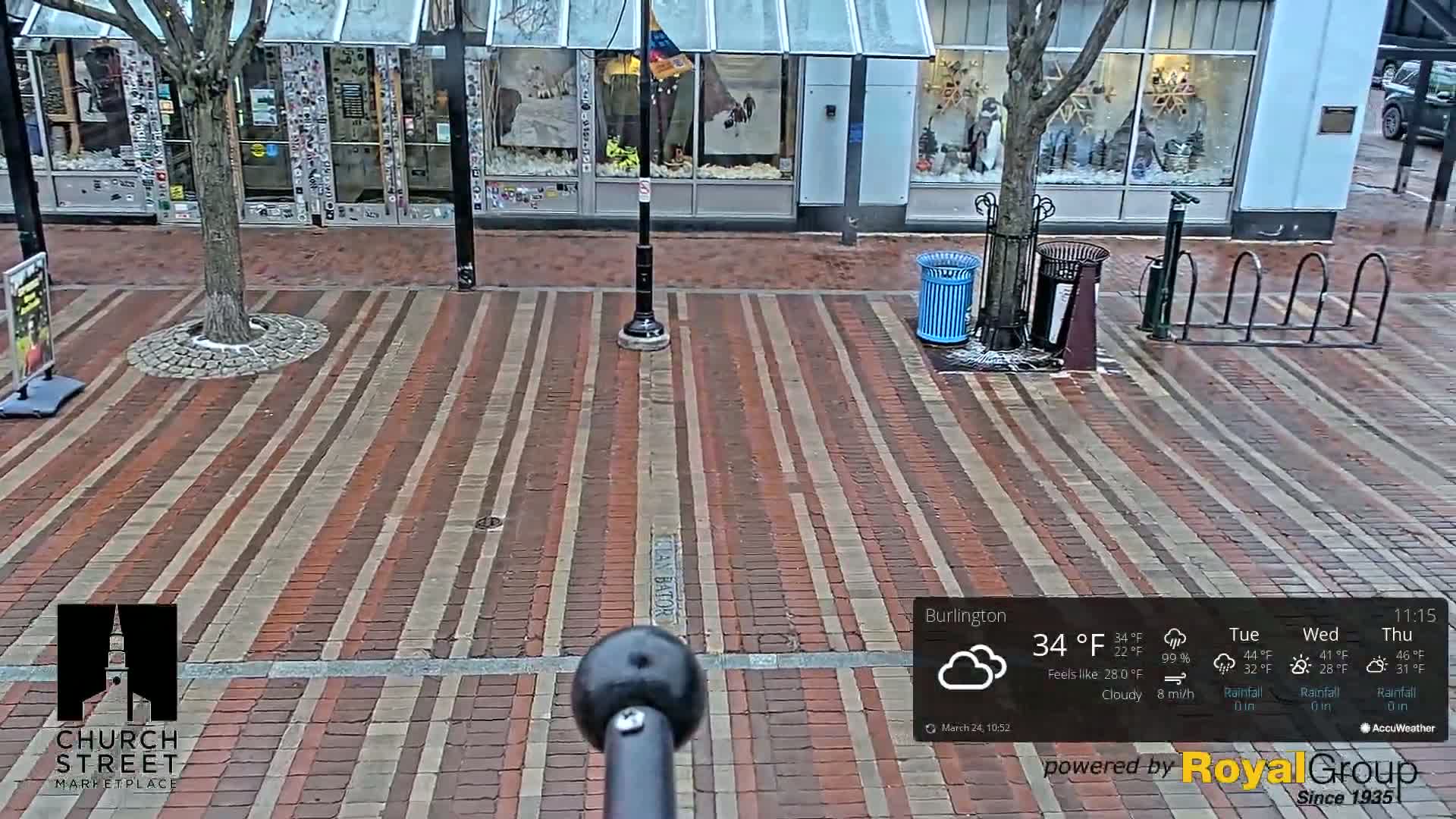 A brick-paved street in front of shops is seen on a cloudy day with a temperature of 34°F.