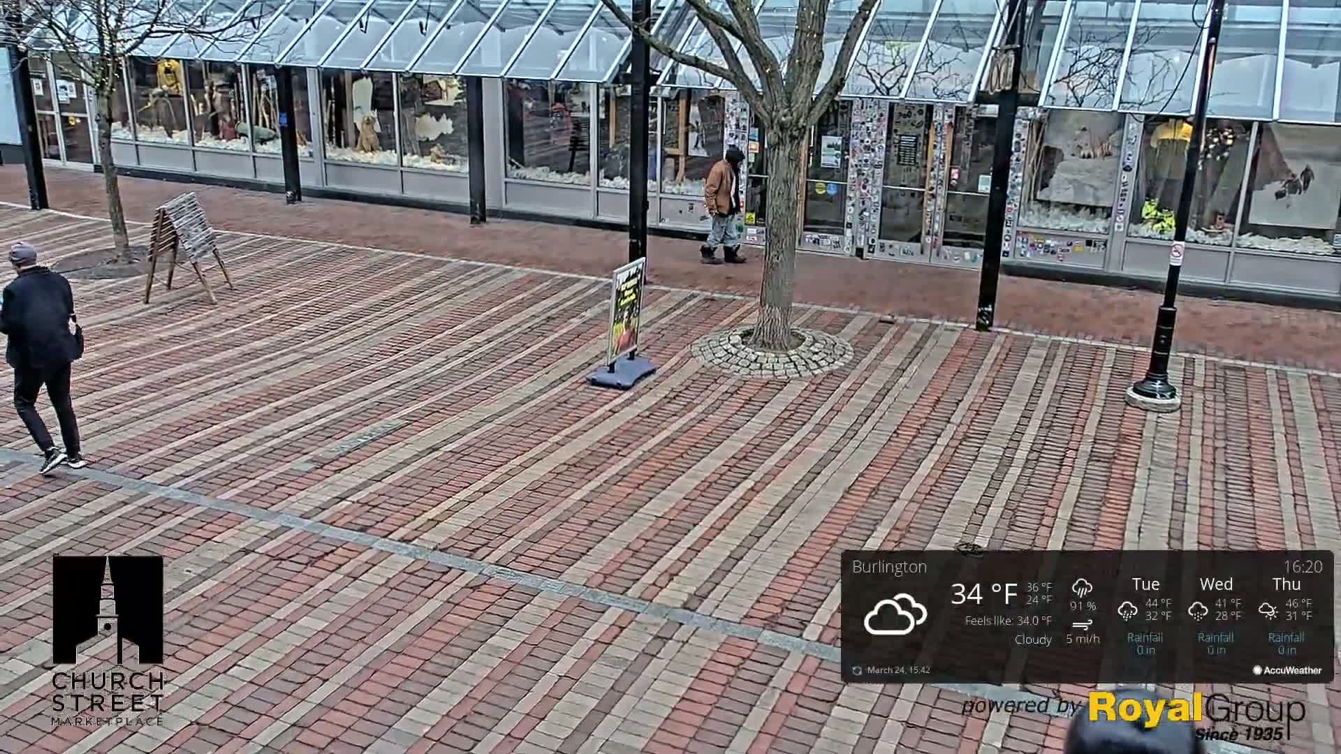 A brick-paved plaza, partially shaded by a tree, is overcast on a cloudy day with two pedestrians walking past storefronts.
