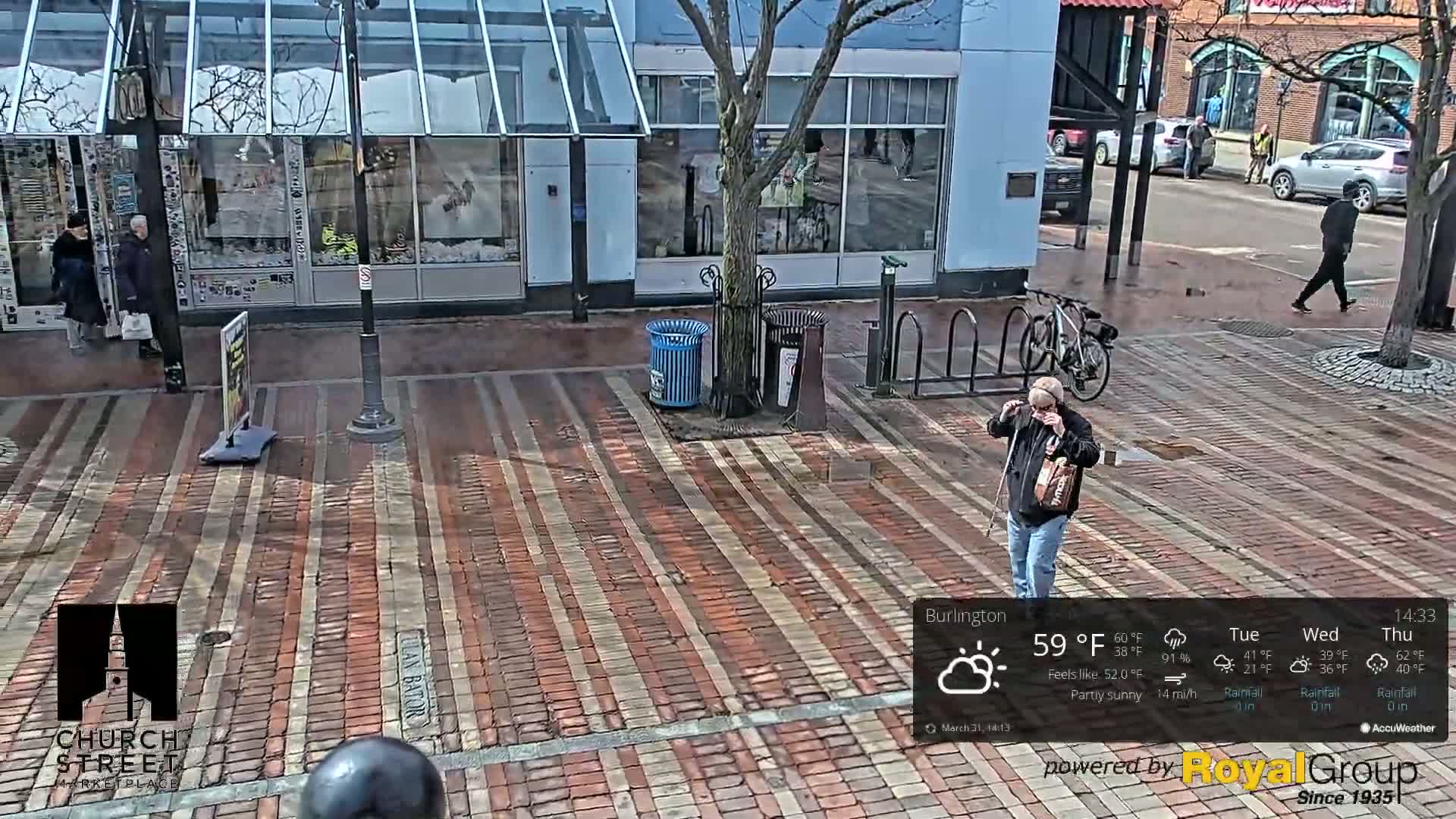 A partly sunny street scene shows pedestrians walking on a brick-paved area in front of shops, with a bicycle rack and trash cans present.
