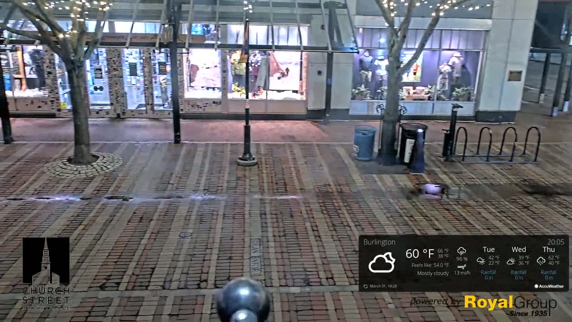 A mostly cloudy night scene shows a brick-paved street in front of shops with mannequins in the windows, a tree in the middle, and a bike rack.