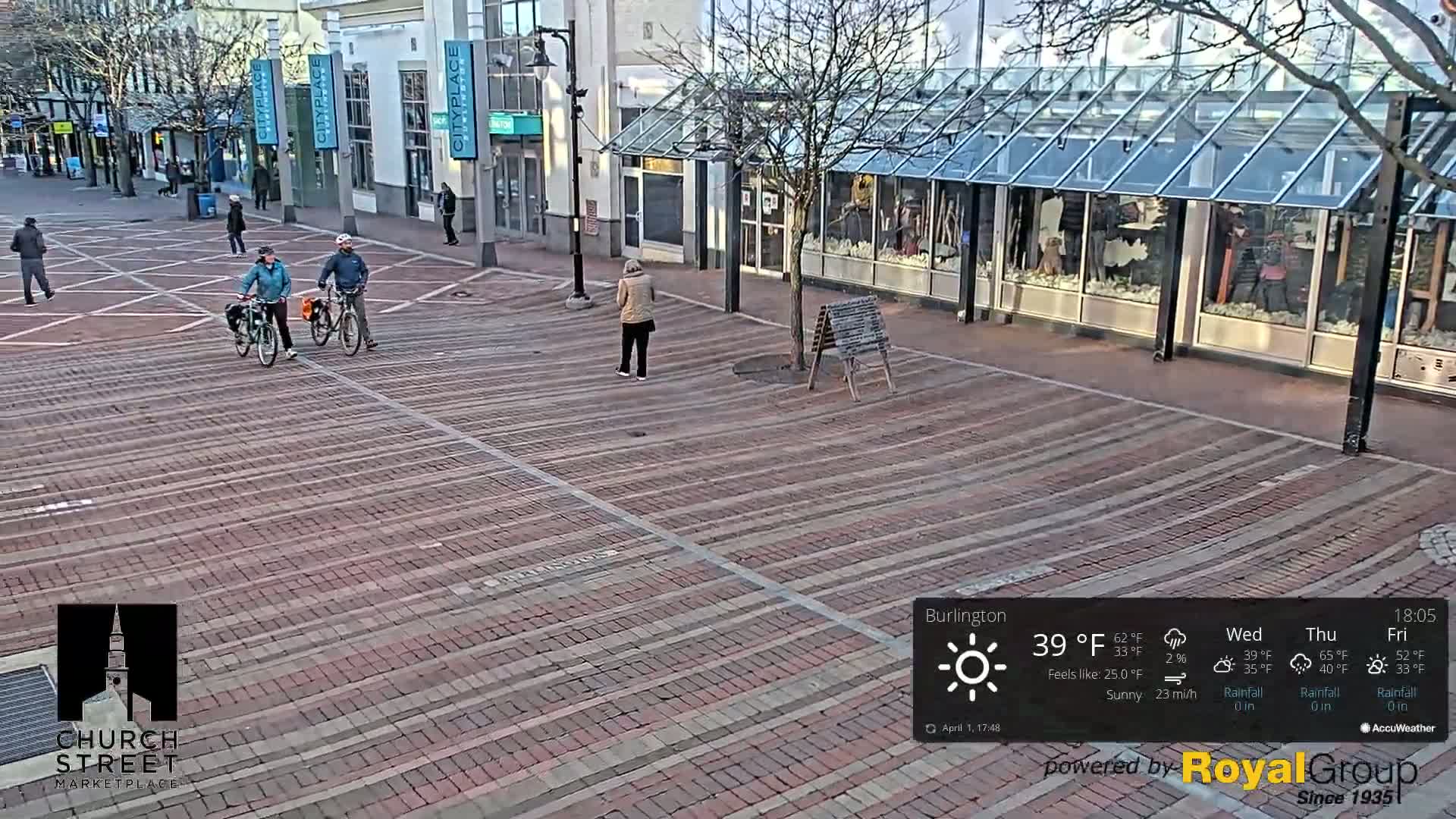 A brick paved city street with a few pedestrians and two cyclists on bicycles under sunny conditions.