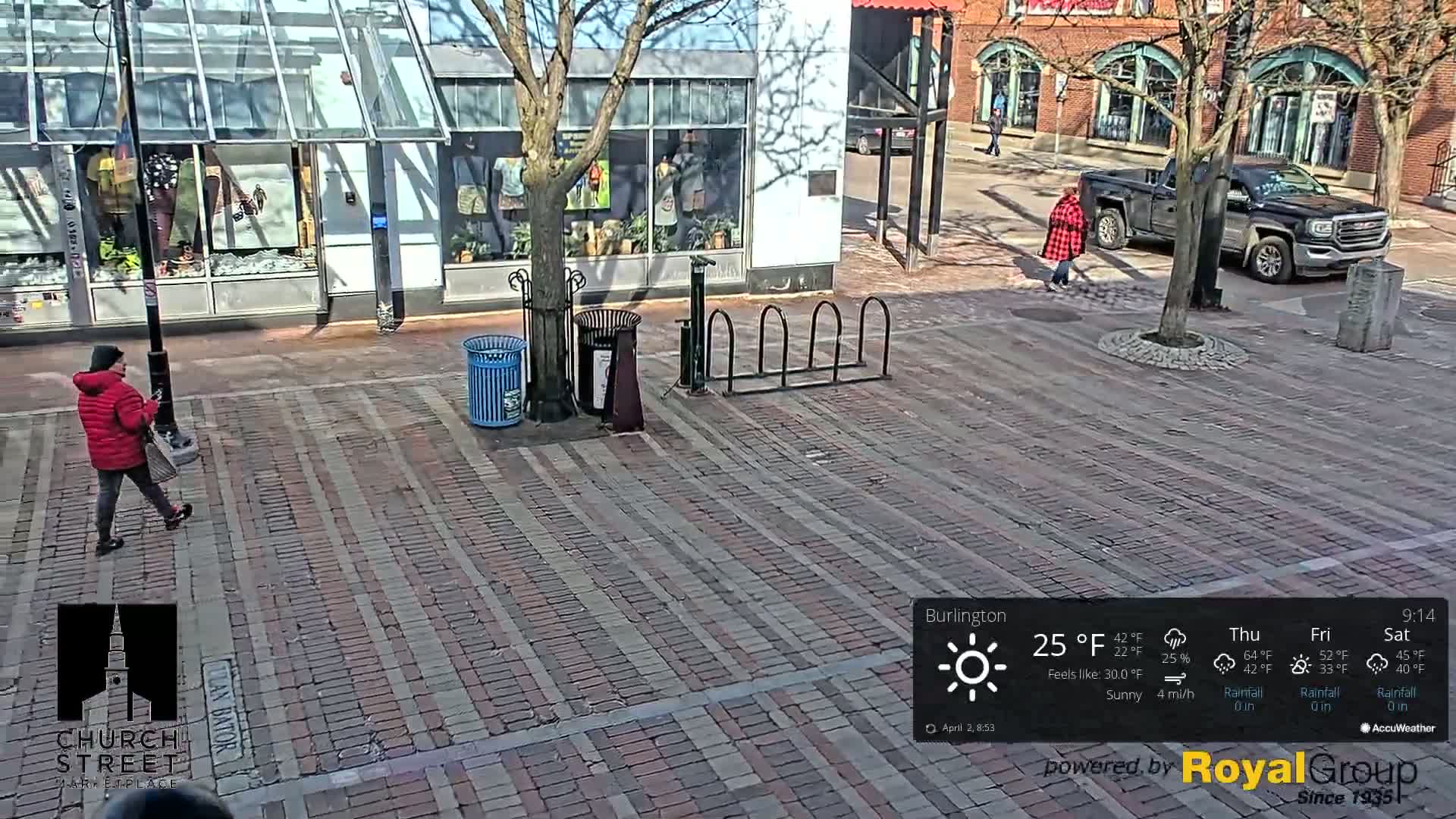 A brick-paved city square is seen on a sunny day with a person in a red jacket walking, a parked truck, and bike racks.