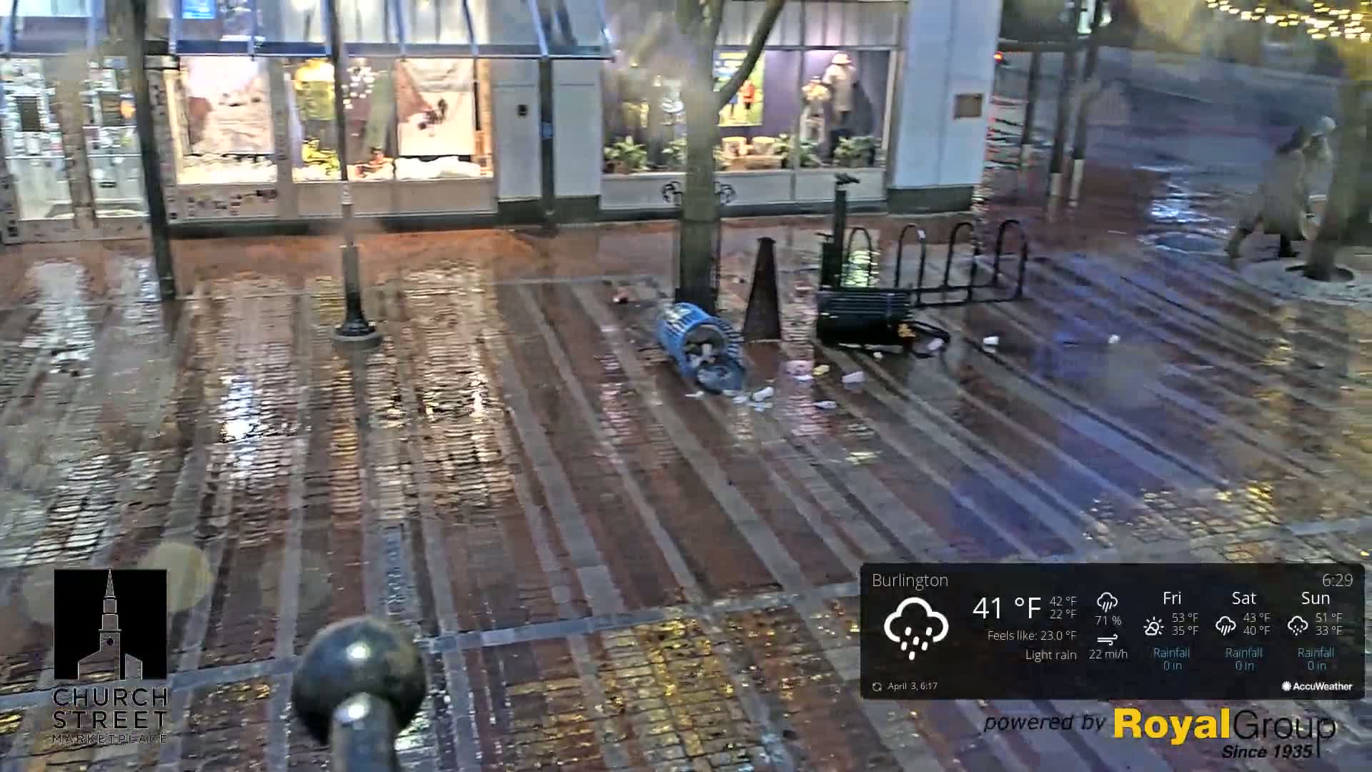 A wet, brick-paved street in front of storefronts is littered with trash near an overturned blue bin, under light rain.
