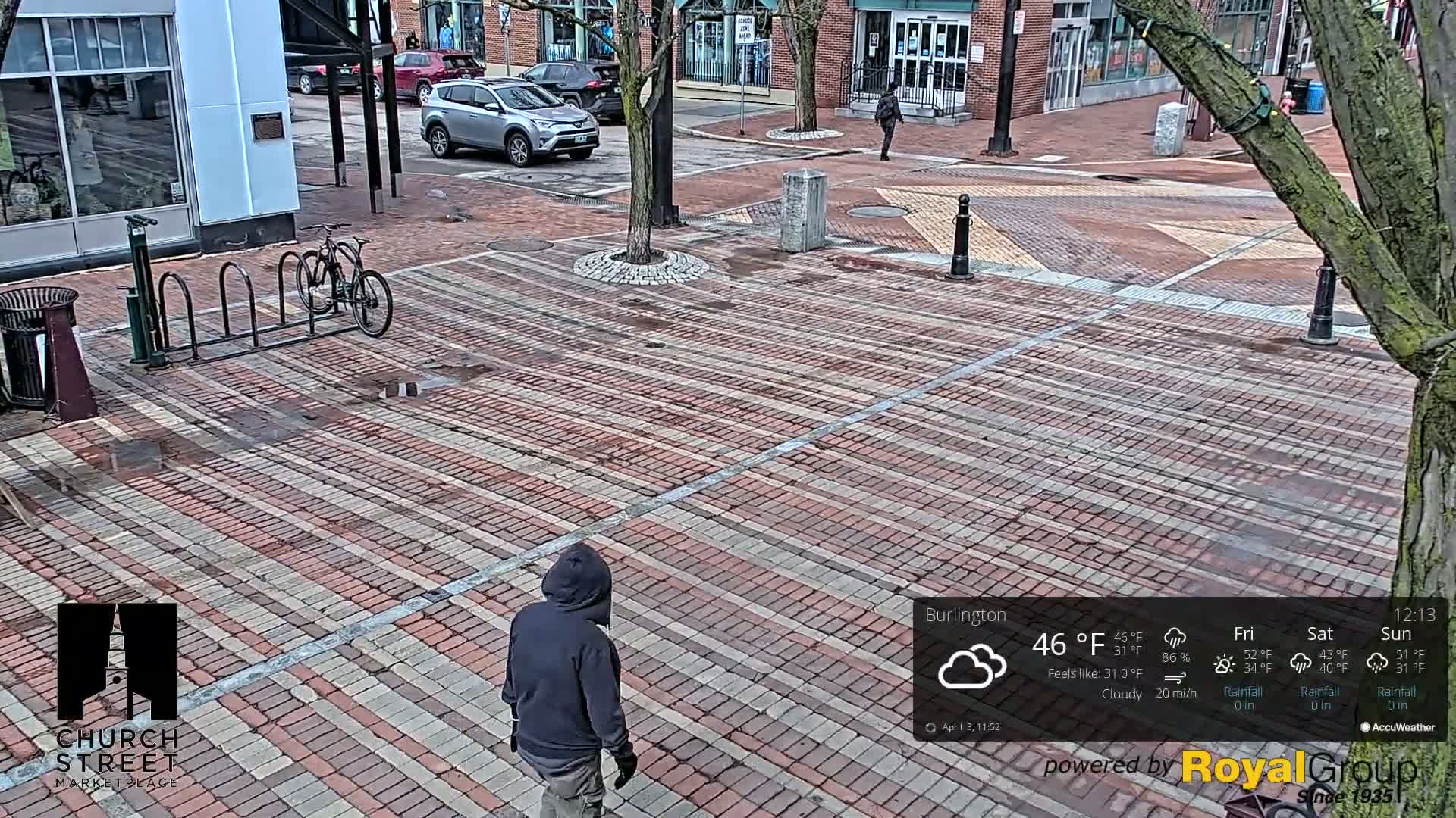A brick plaza, wet from recent rain, shows a person walking, a bicycle in a rack, and cars parked along the street.
