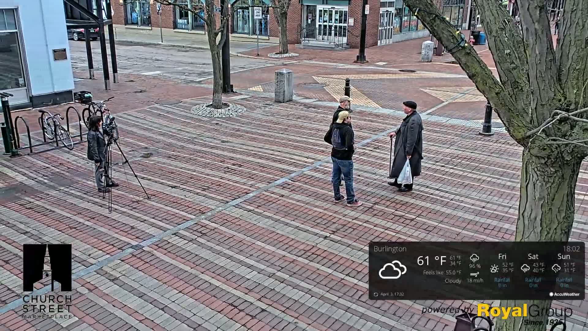 A person films three people conversing on a brick paved street under cloudy skies.
