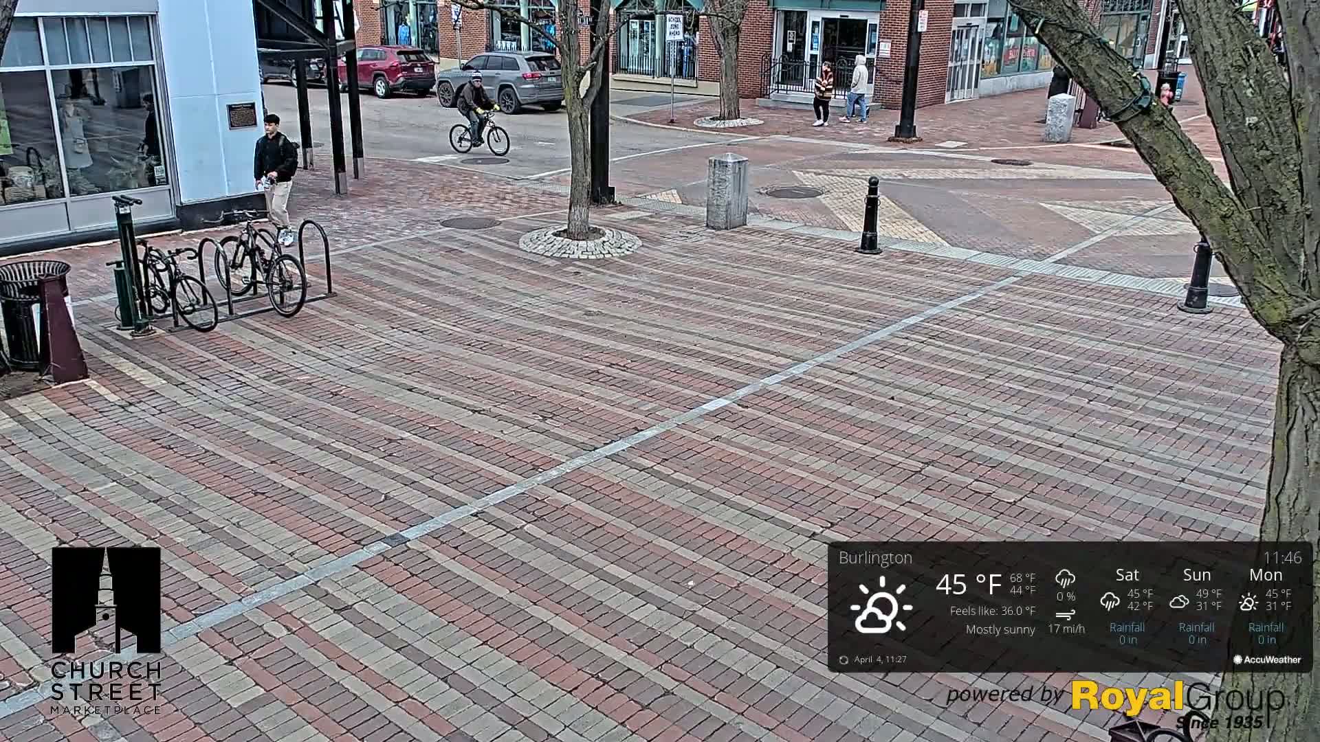 A mostly sunny brick-paved street scene shows a few pedestrians and bicyclists, with parked bicycles at a bike rack, under a partly sunny sky at 45°F.