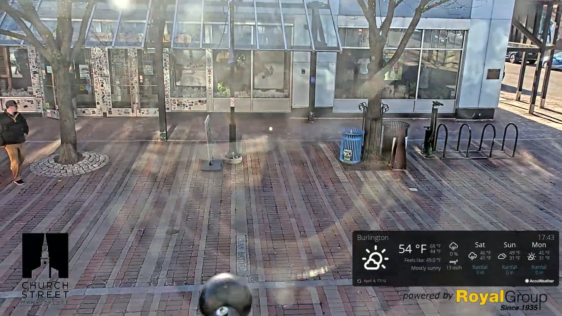 A mostly sunny brick paved area with a person walking near a building, bike racks, and trash cans.