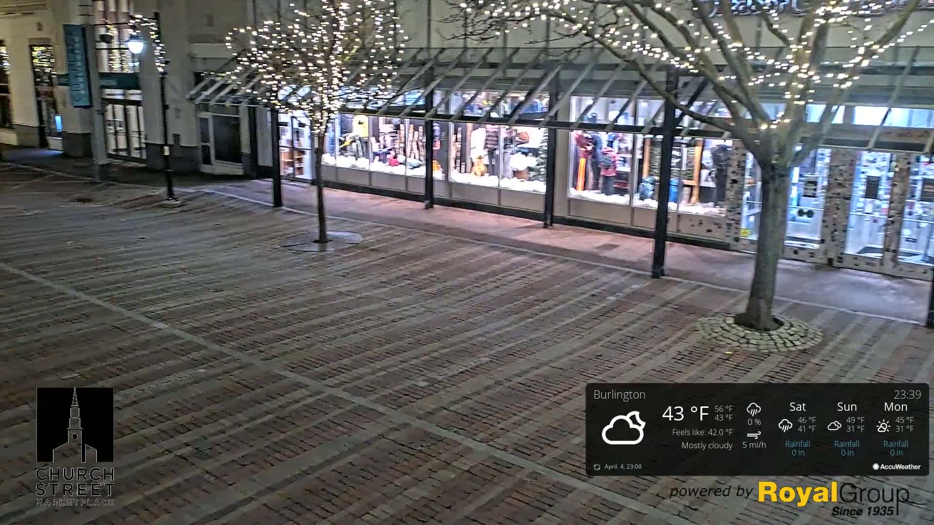 A nighttime view of a brick paved street in front of a store with illuminated windows and a tree adorned with string lights under mostly cloudy skies.