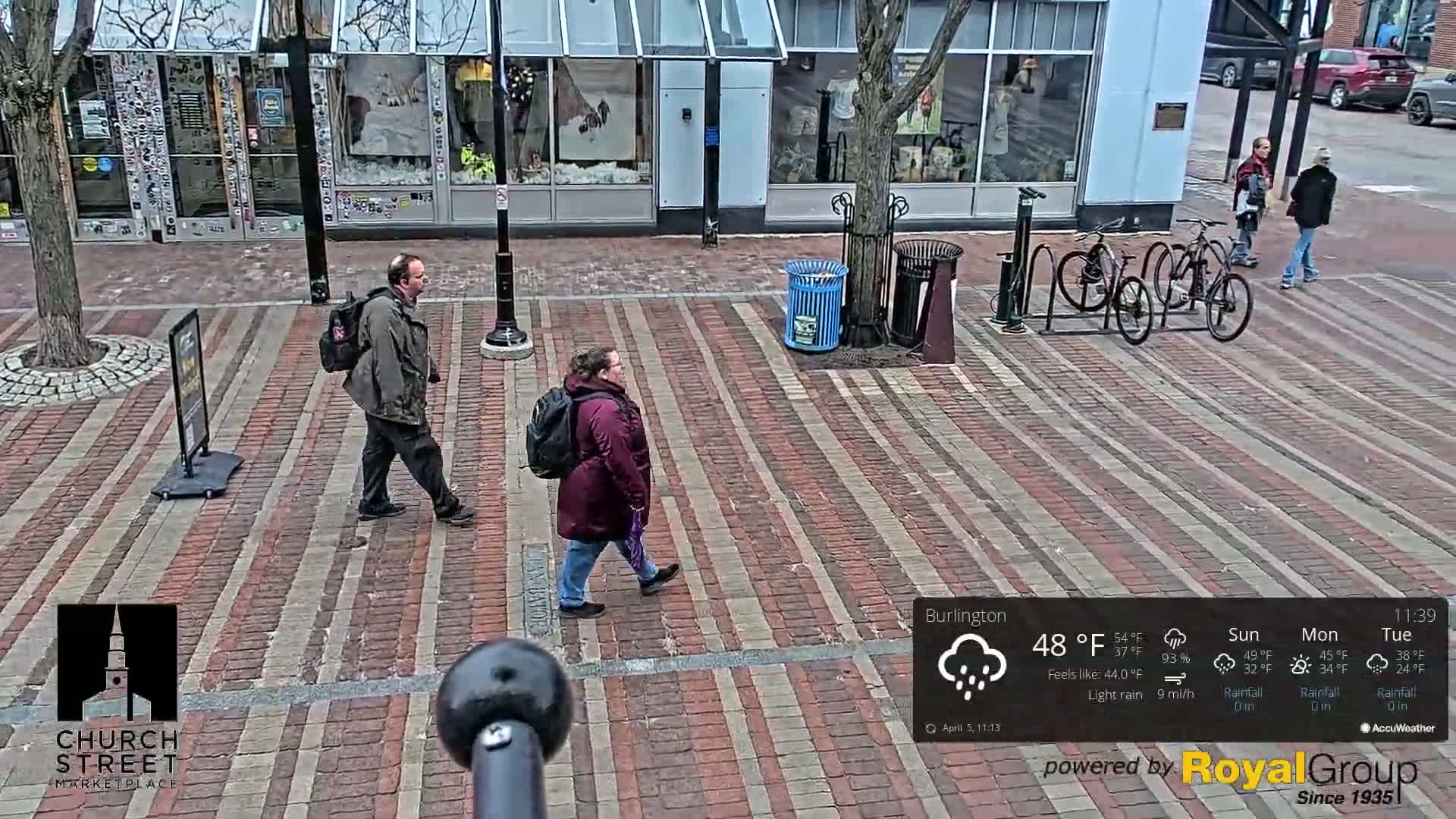 Three people walk on a brick-paved street in front of storefronts under light rain.