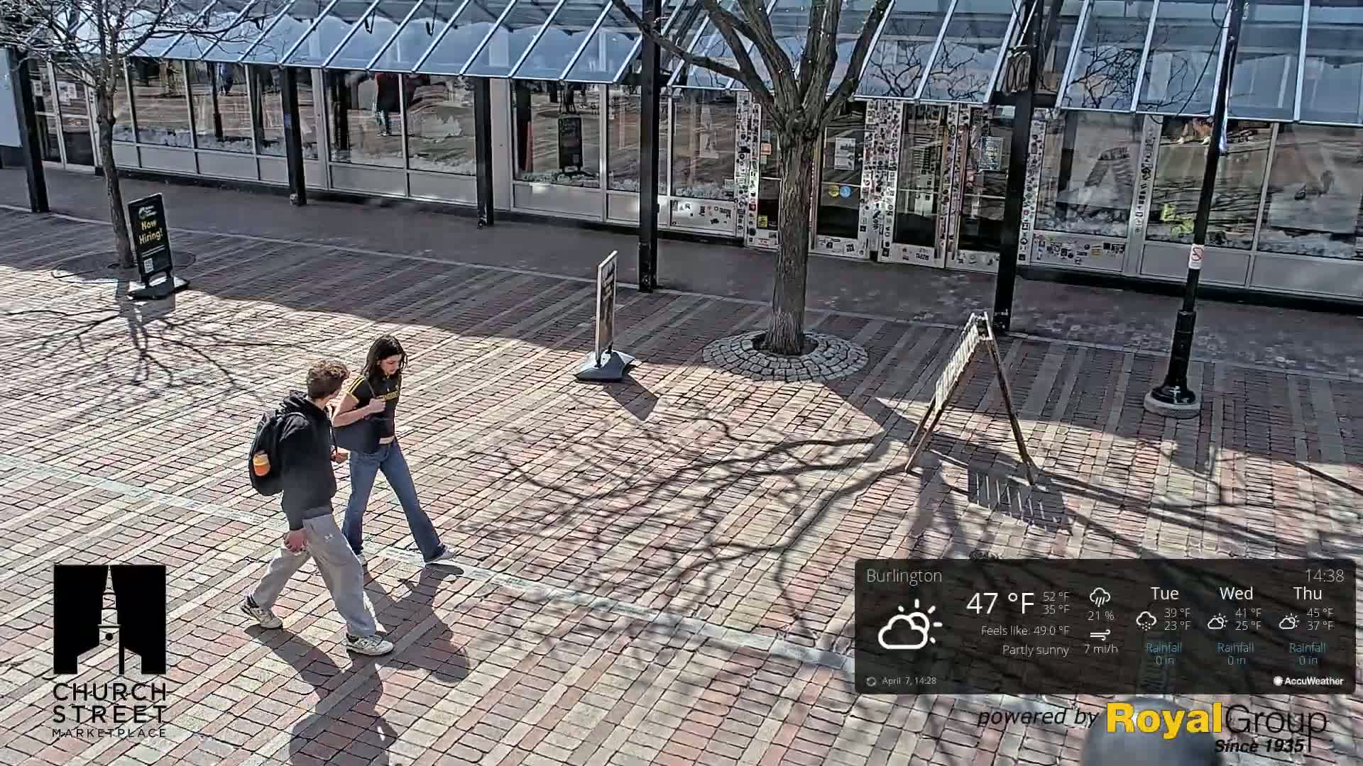 Two people walk across a brick plaza in partly sunny weather, passing in front of a building with large glass windows.