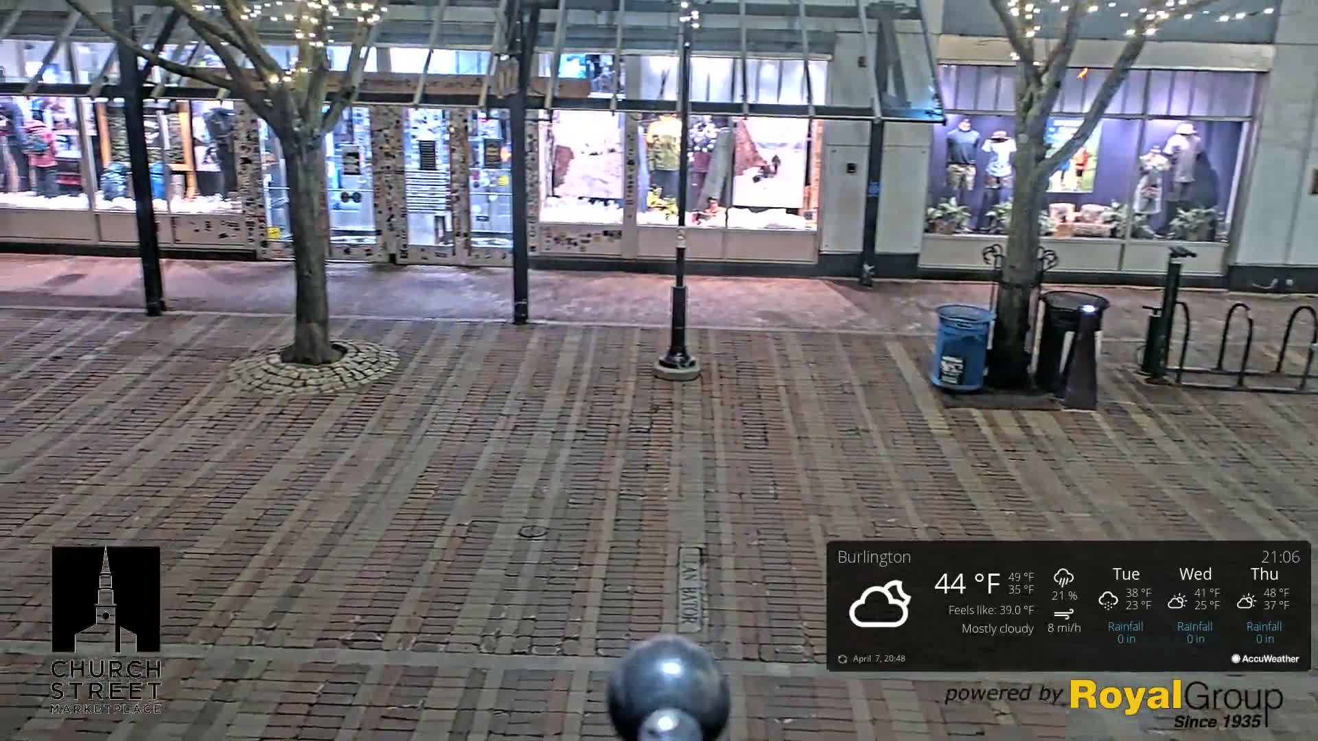 A mostly cloudy night scene shows a brick-paved street in front of storefronts with some trees and bike racks, under string lights.