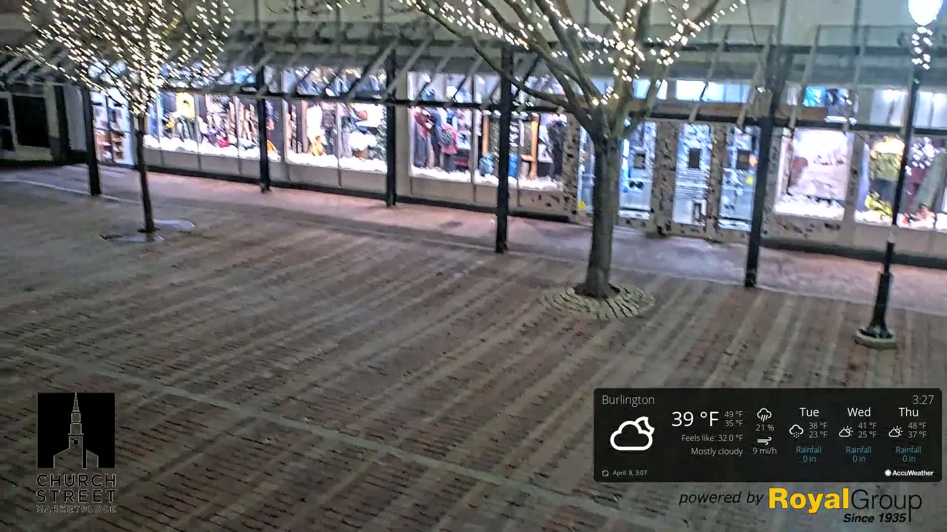 A mostly cloudy night scene shows a brick plaza in front of a row of storefronts decorated with string lights.