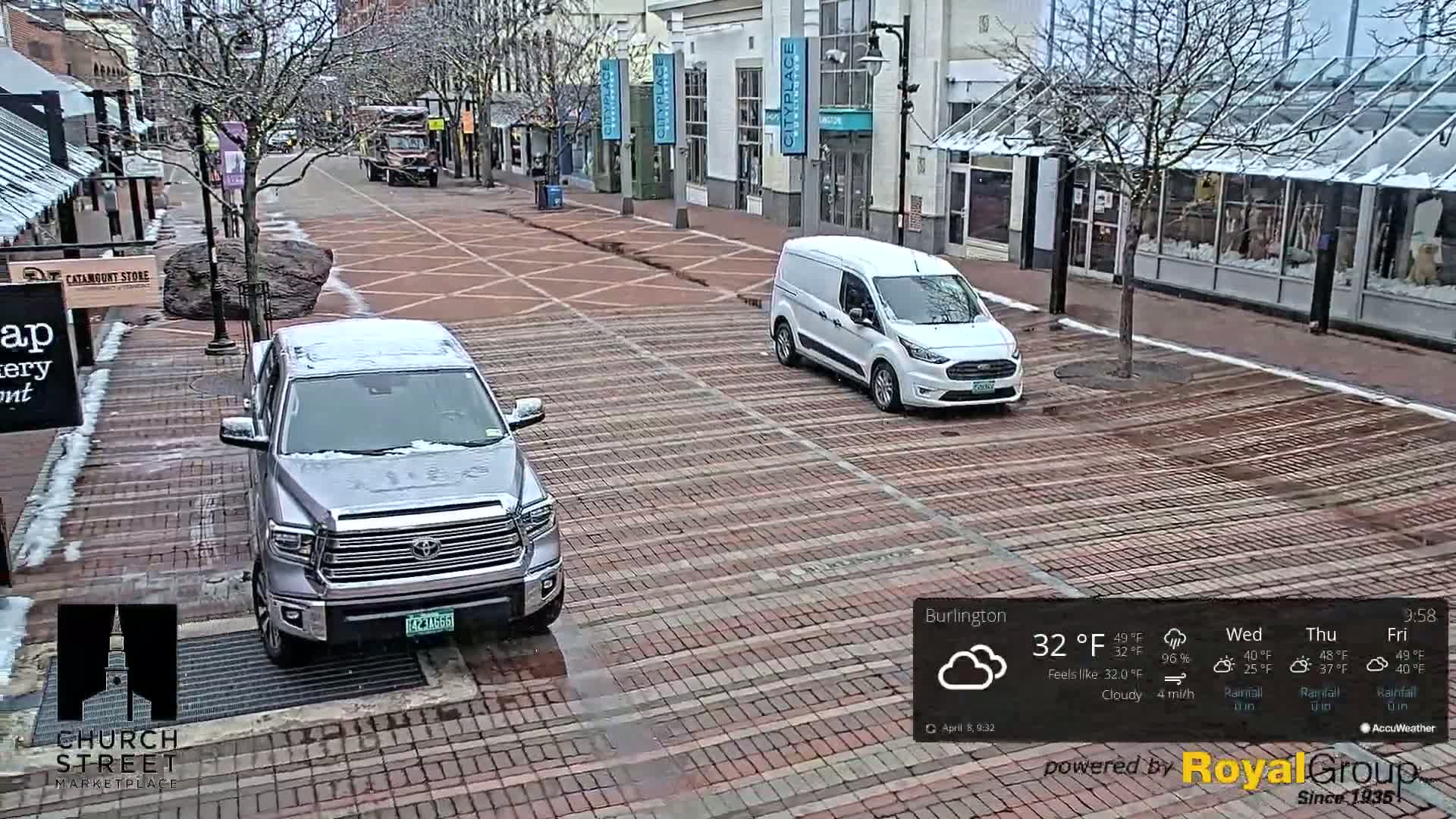 A brick-paved street with a silver pickup truck and a white van parked, lightly dusted with snow under cloudy skies.