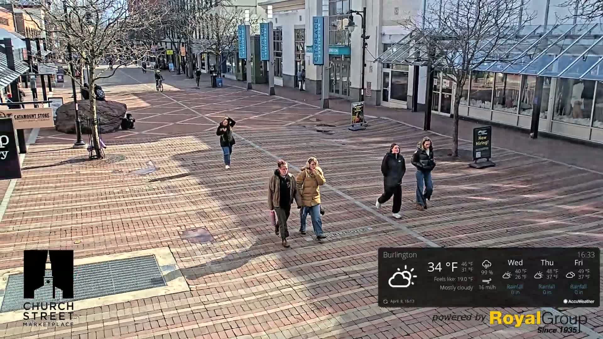 A mostly cloudy, sunny brick-paved pedestrian area with several people walking, surrounded by buildings.