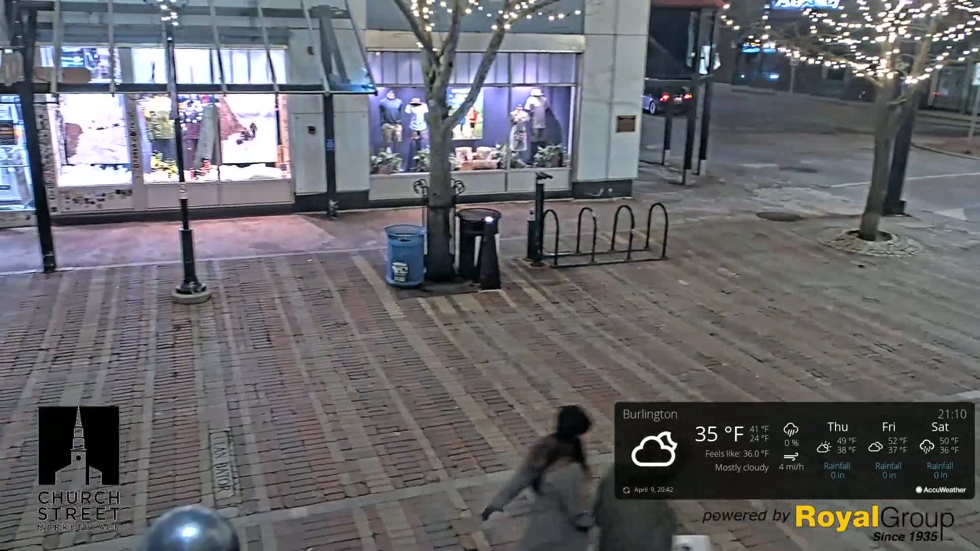 A mostly cloudy night scene shows a brick-paved street with a person walking past a bike rack and store windows.