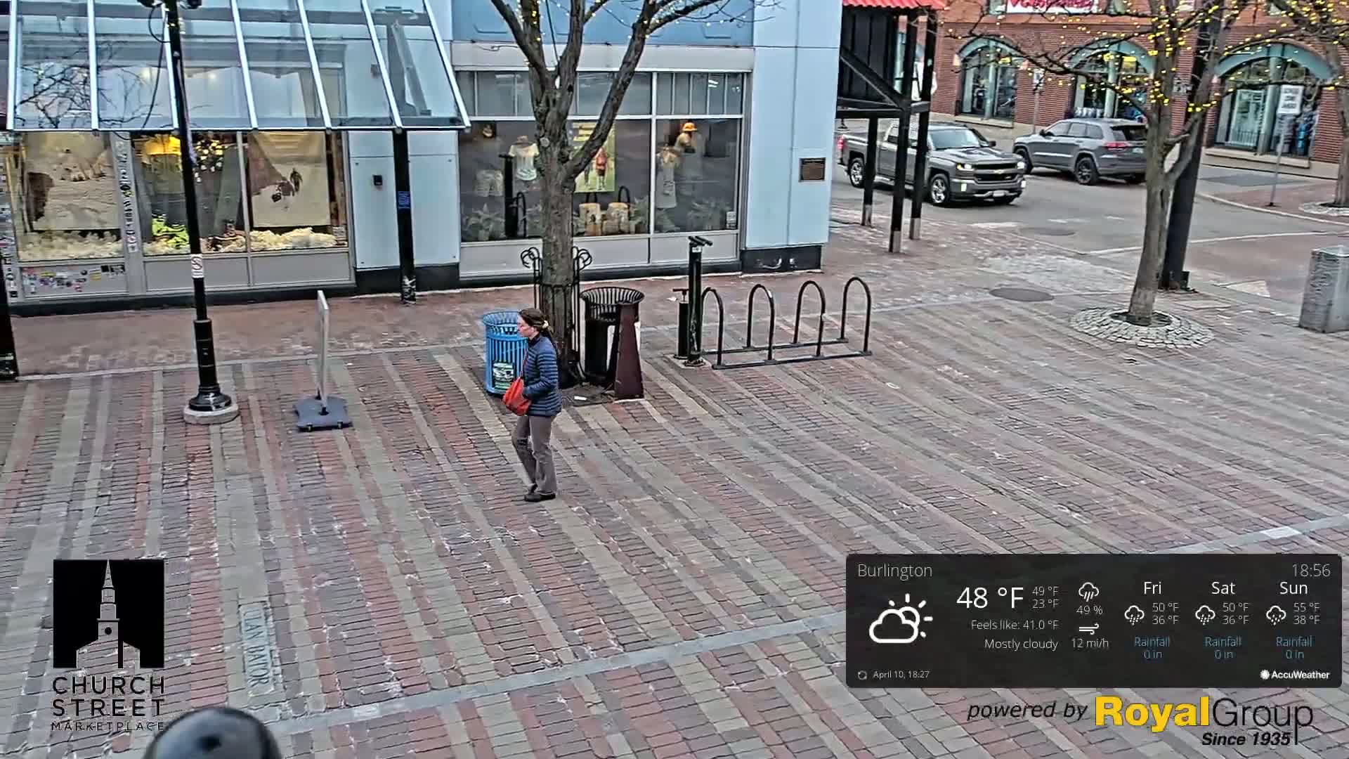 A woman walks across a brick-paved street in front of shops under a mostly cloudy sky.