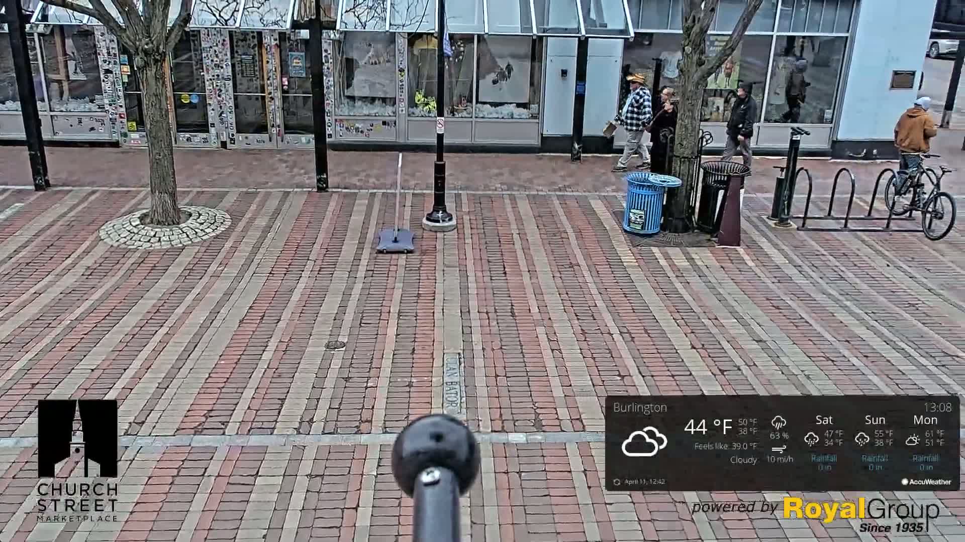 A brick-paved city street scene on a cloudy day shows several pedestrians, a bicycle leaning against a rack, and a small tree in a circular stone border.