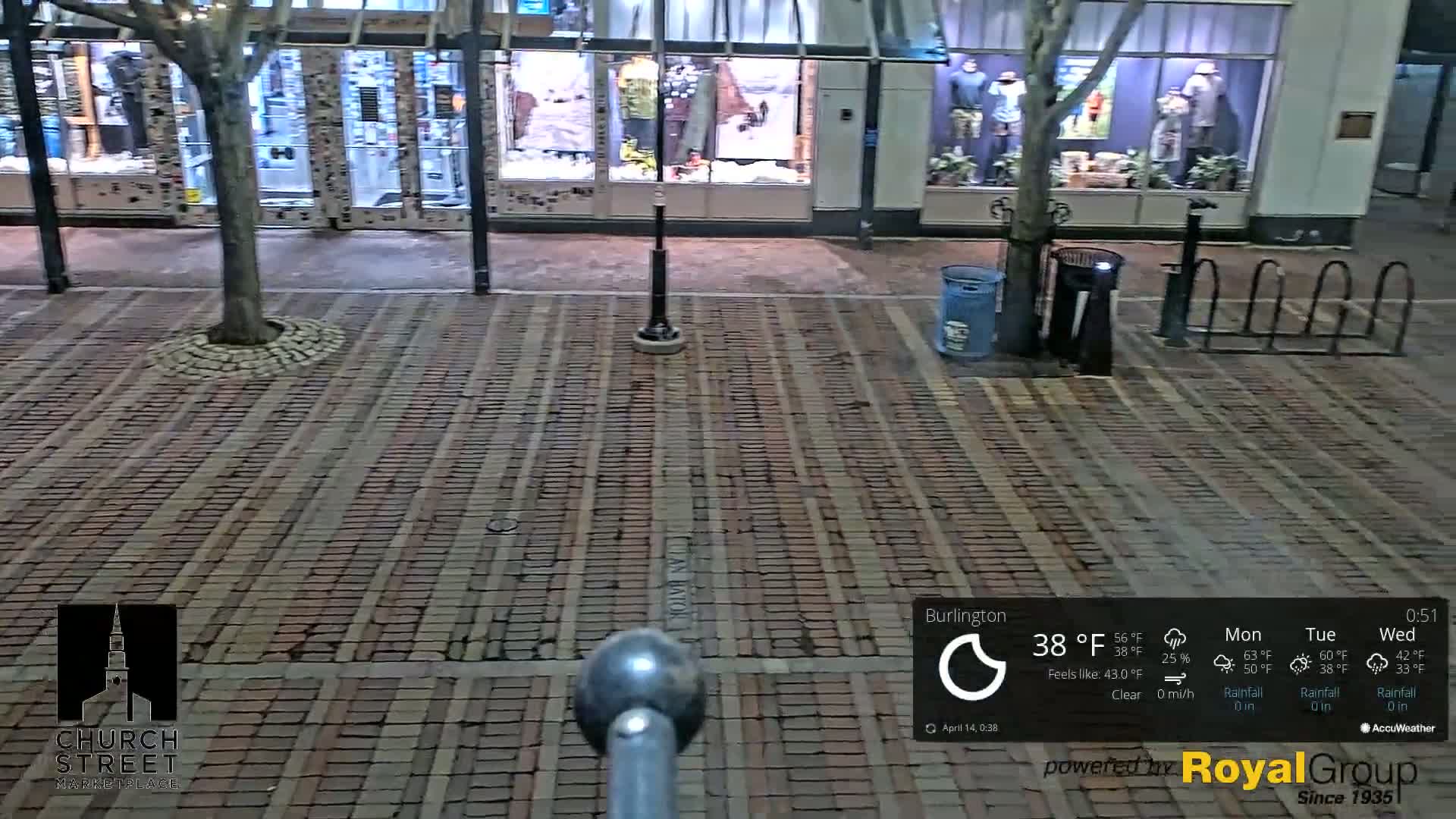 A brick paved street at night, under clear skies with a temperature of 38°F, shows the fronts of stores with mannequins in their windows, a small tree, trash cans, and bike racks.