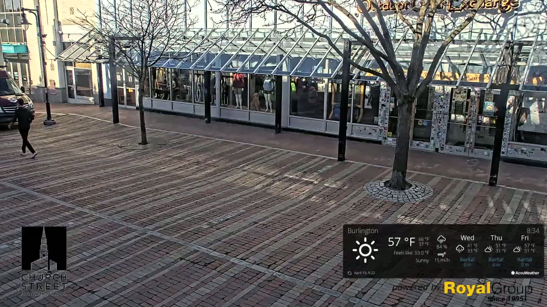 A person walks across a brick-paved plaza in front of a glass-fronted building on a sunny day with a 15 mi/h wind.