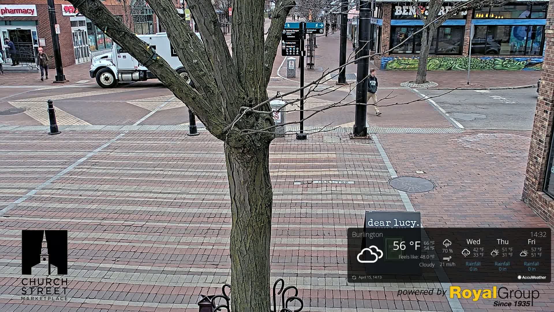 A brick-paved street scene on a cloudy day shows a tree in the foreground, a person walking, and a white truck parked nearby.