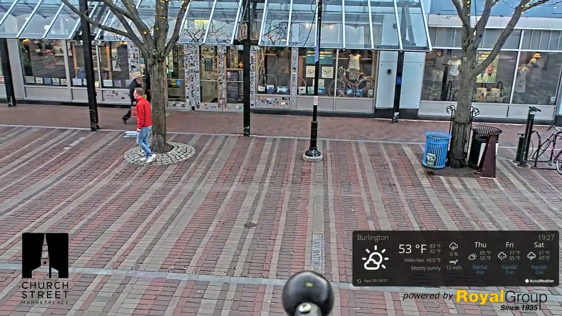 A mostly sunny street scene shows two people walking on a brick-paved walkway in front of shops with glass awnings.