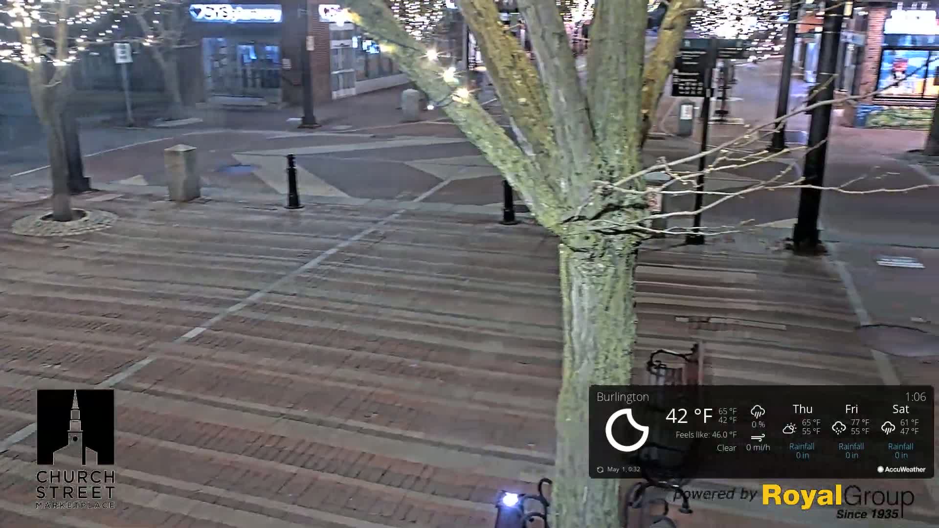 A brick-paved plaza at night, illuminated by streetlights and string lights on a tree, is clear and 42°F.