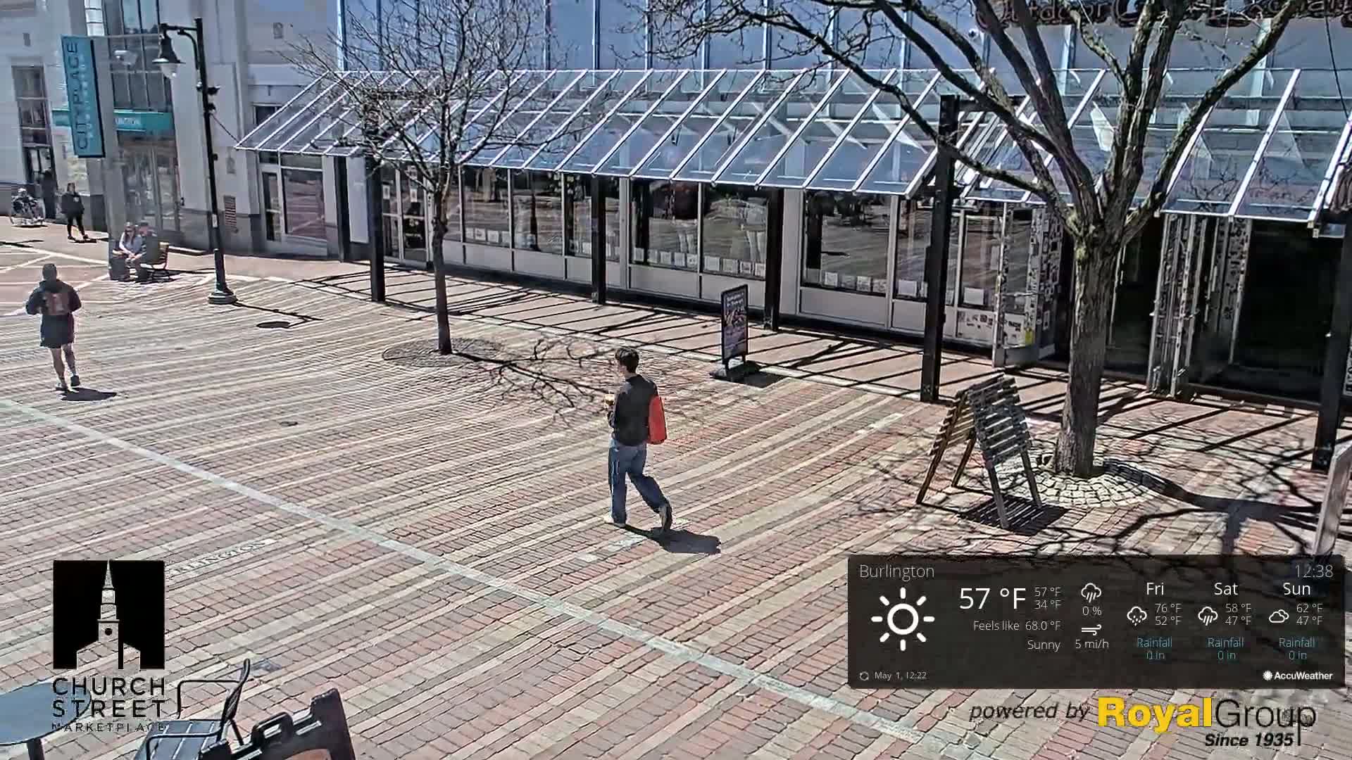 A brick plaza in sunny weather with a few pedestrians and a glass-roofed building in the background.
