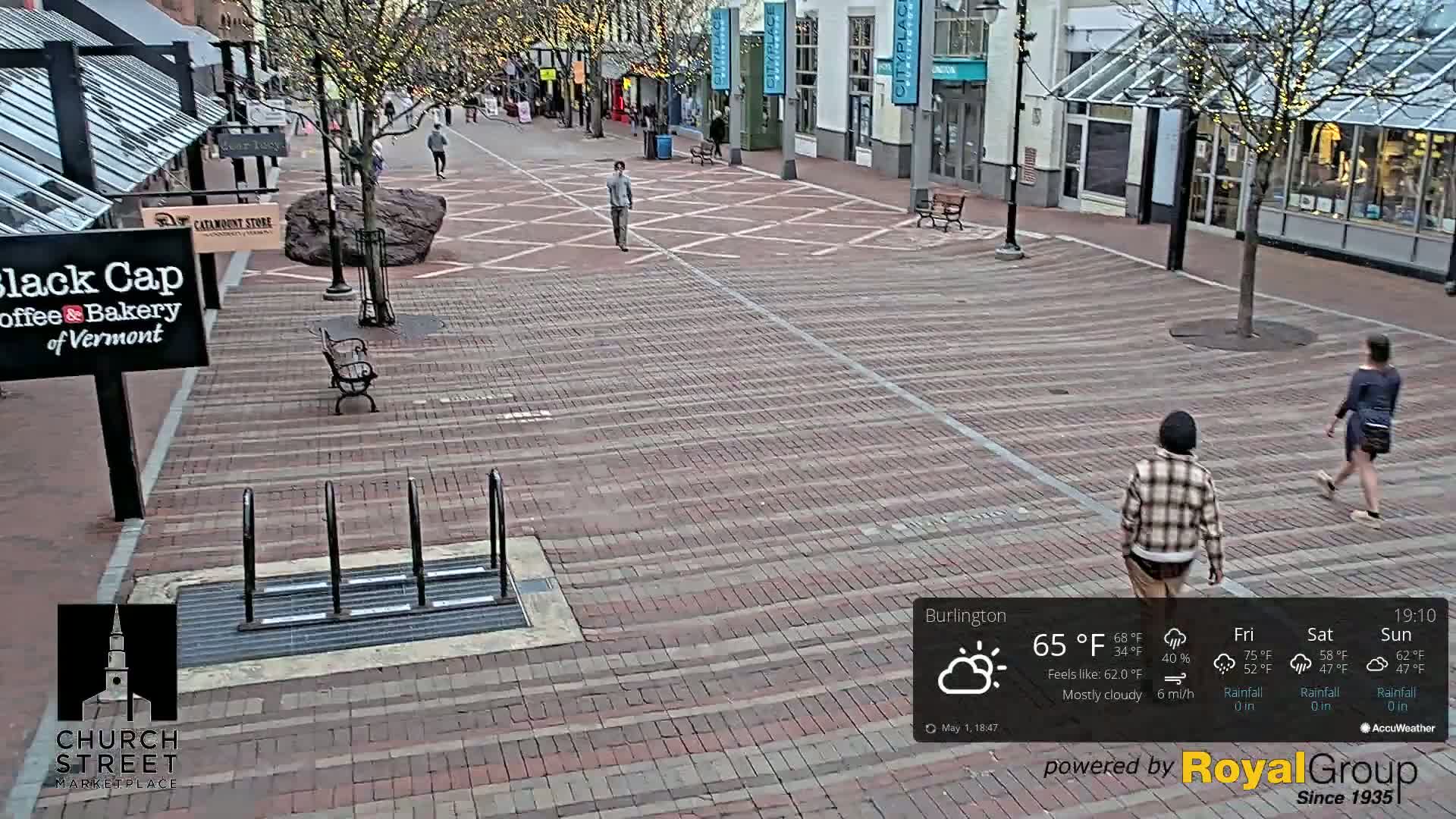 A mostly cloudy brick pedestrian mall with a few people walking, a large rock, and a bicycle rack.