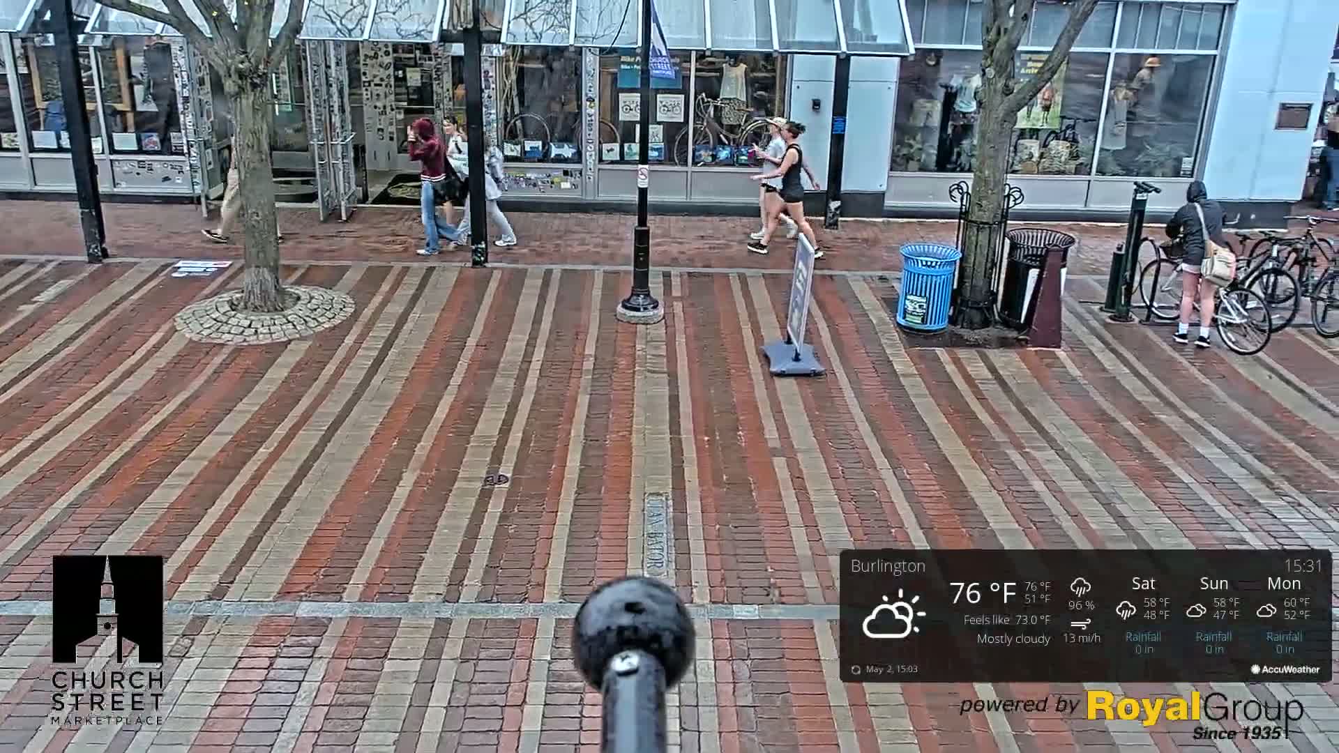 A brick-paved street with pedestrians and bicycles is seen on a mostly cloudy day with a temperature of 76°F and a 13 mph wind.