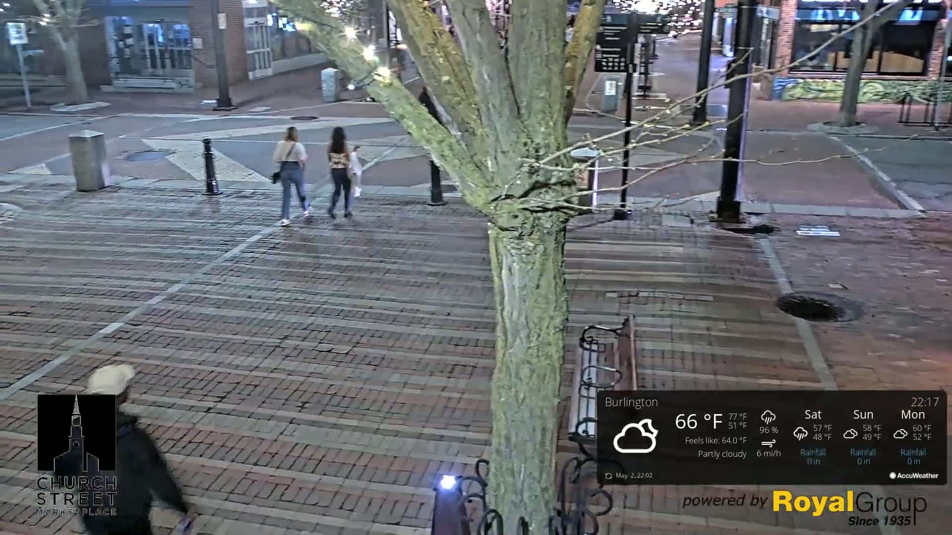 Three people walk on a brick-paved street at night under partly cloudy skies.