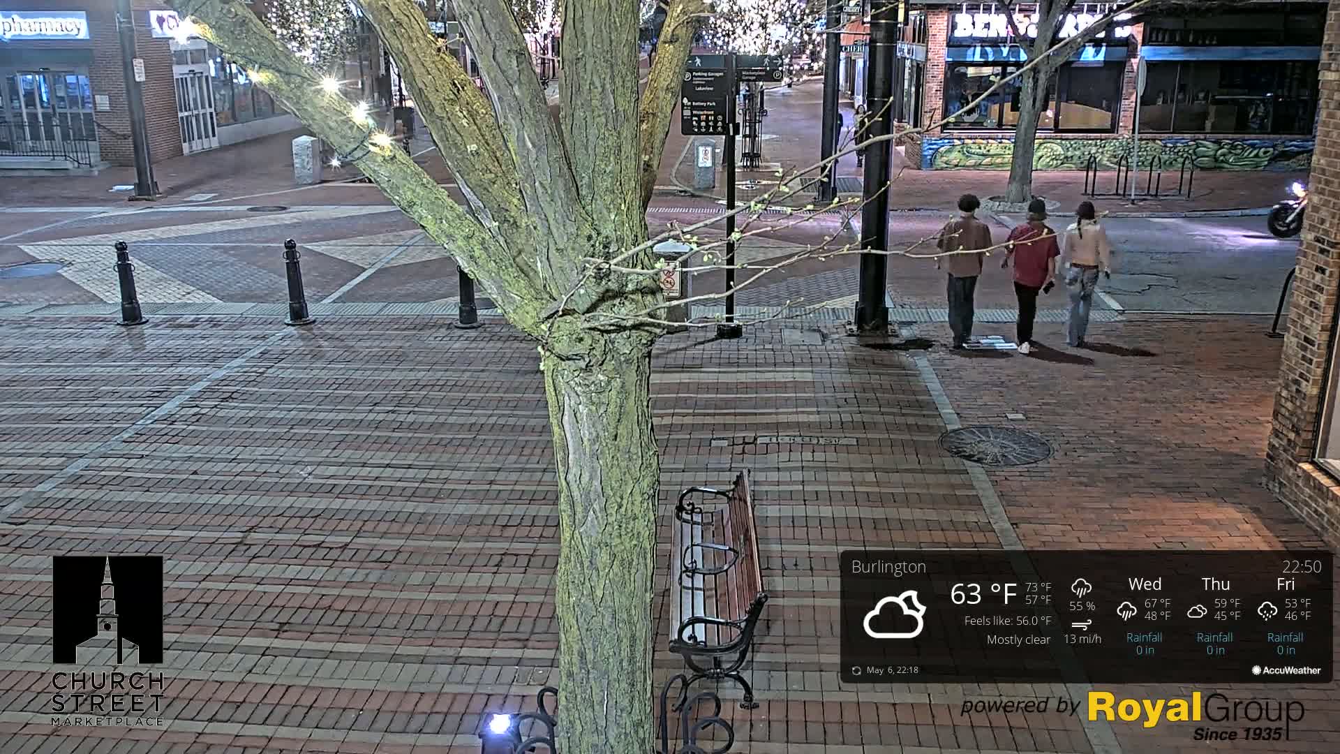 At night, under mostly clear skies, three people walk down a brick-paved street lined with buildings and a tree with string lights.