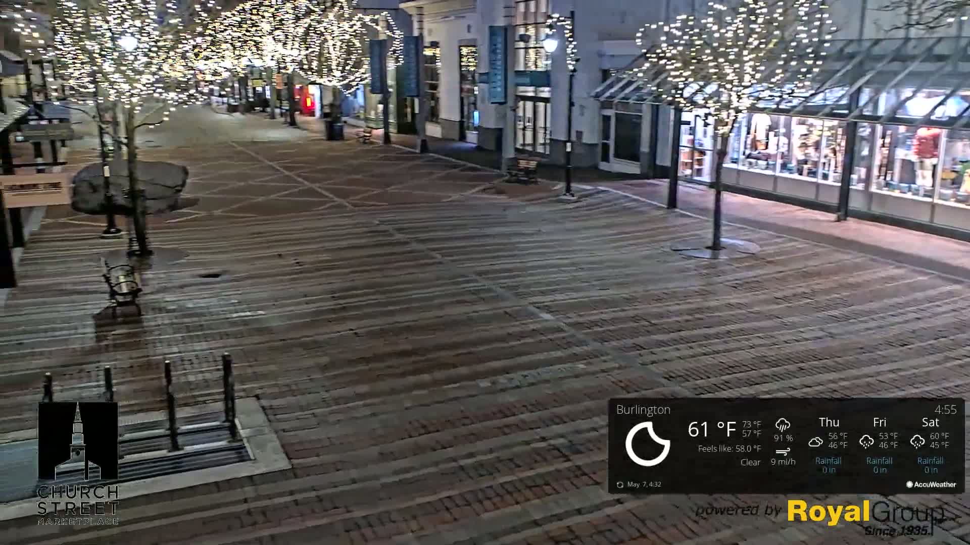 A brick pedestrian street at night is illuminated by string lights on trees and the lights of nearby buildings, under clear skies.