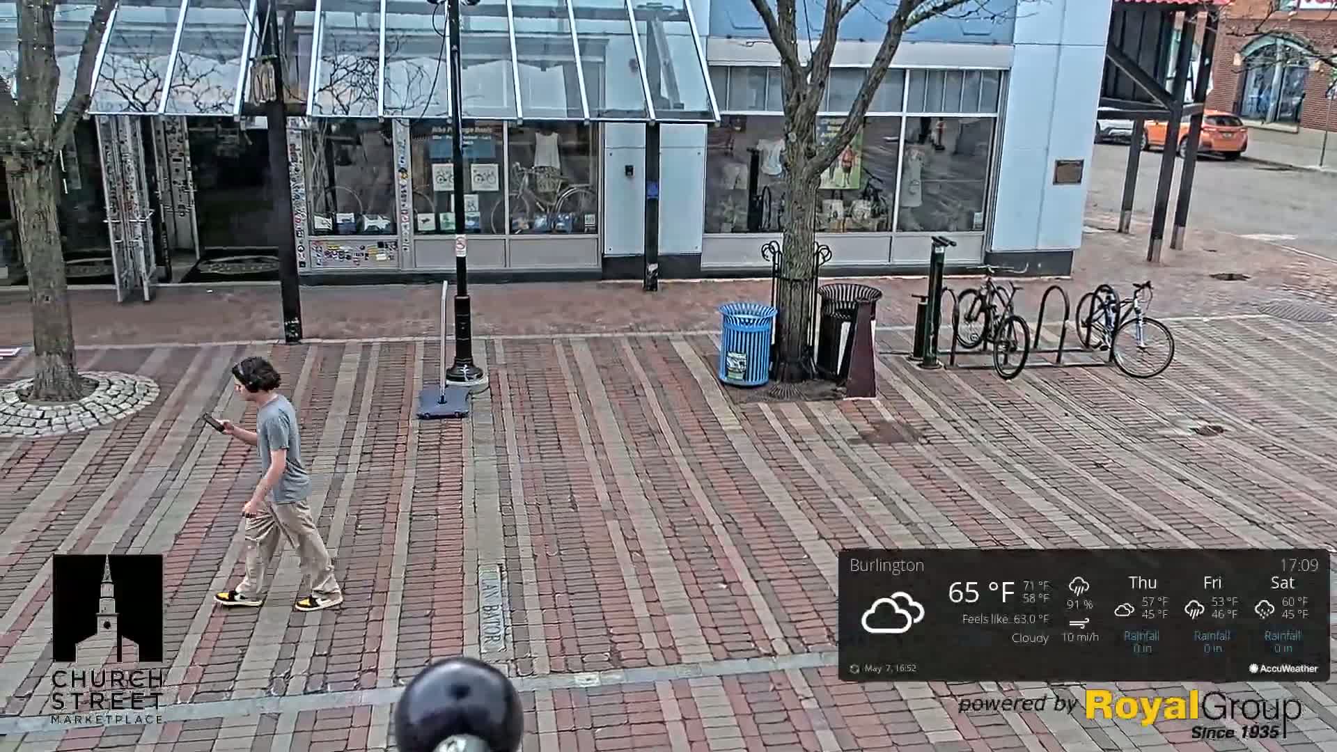 A person walks on a brick-paved street past a bike rack under a cloudy sky.