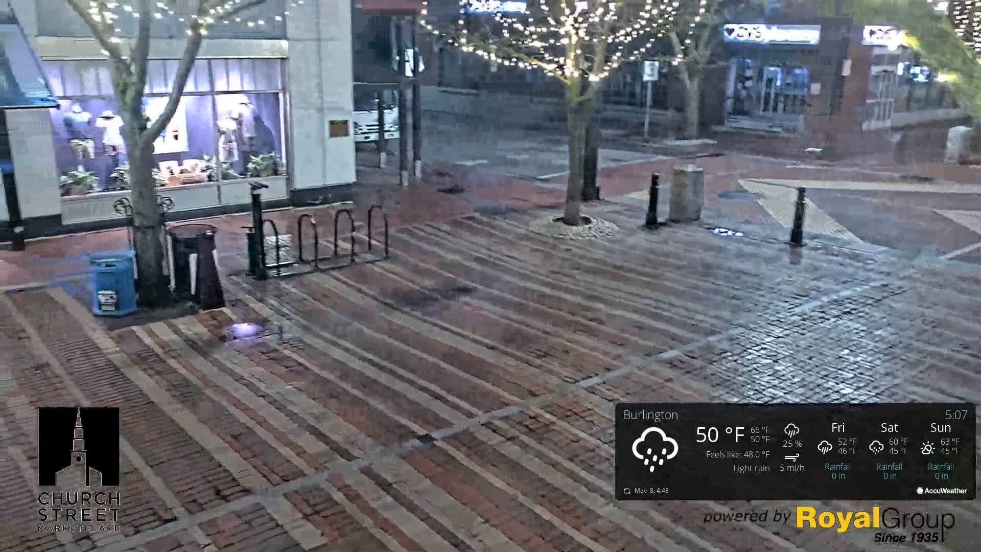 A wet brick plaza is seen at night under light rain, with a tree and bicycle rack near a storefront decorated with string lights.