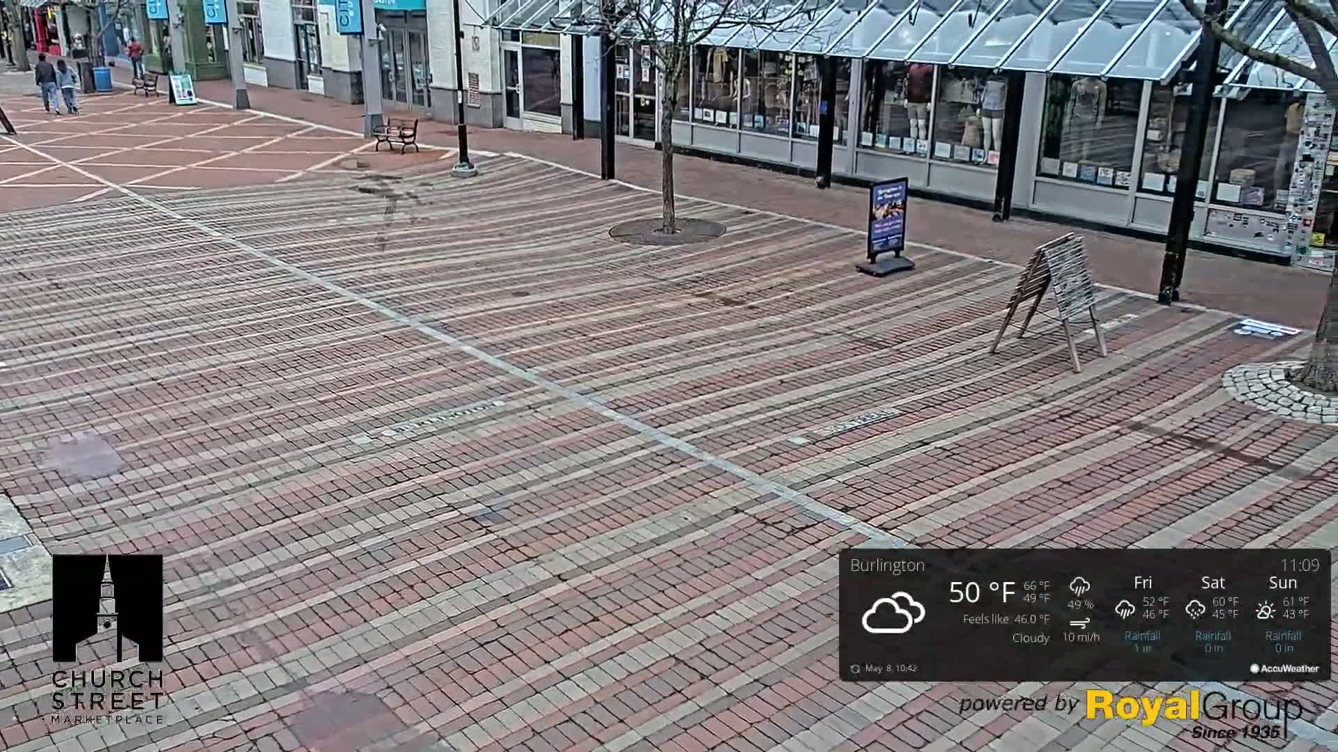 A brick-paved plaza, under cloudy skies, is mostly empty except for a few pedestrians and some street furniture.