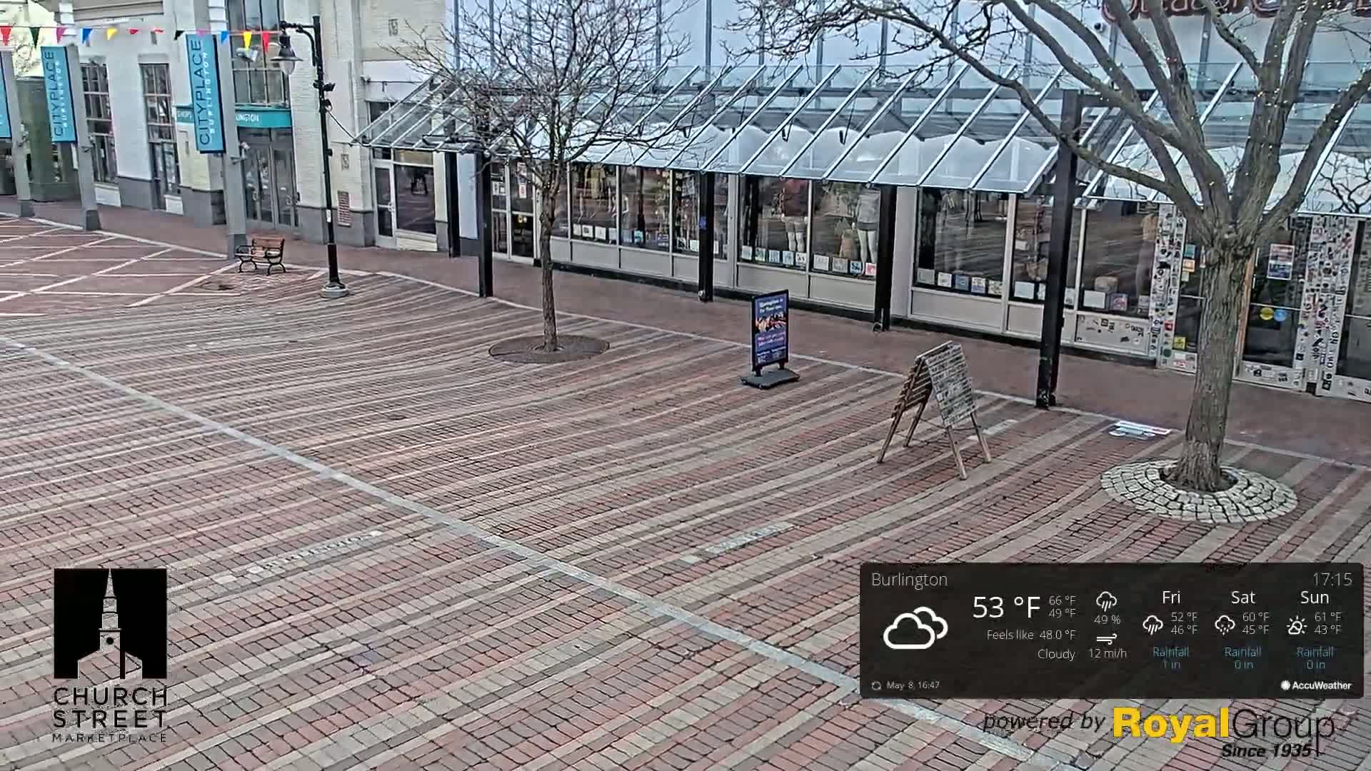A brick-paved plaza sits in front of several shops under a cloudy sky, with a lone bench and a tree present.