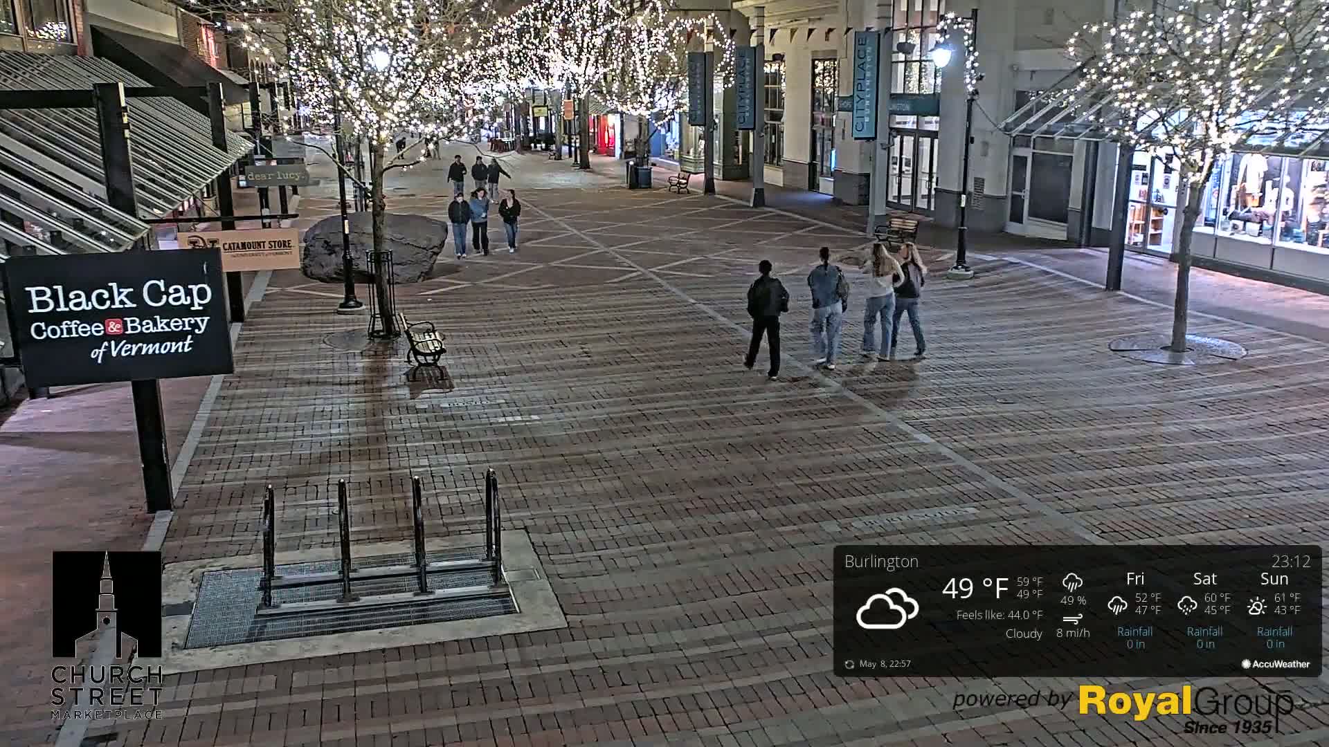 A brick pedestrian area at night is lit by string lights on trees, with several people walking and small groups standing; the weather is cloudy.