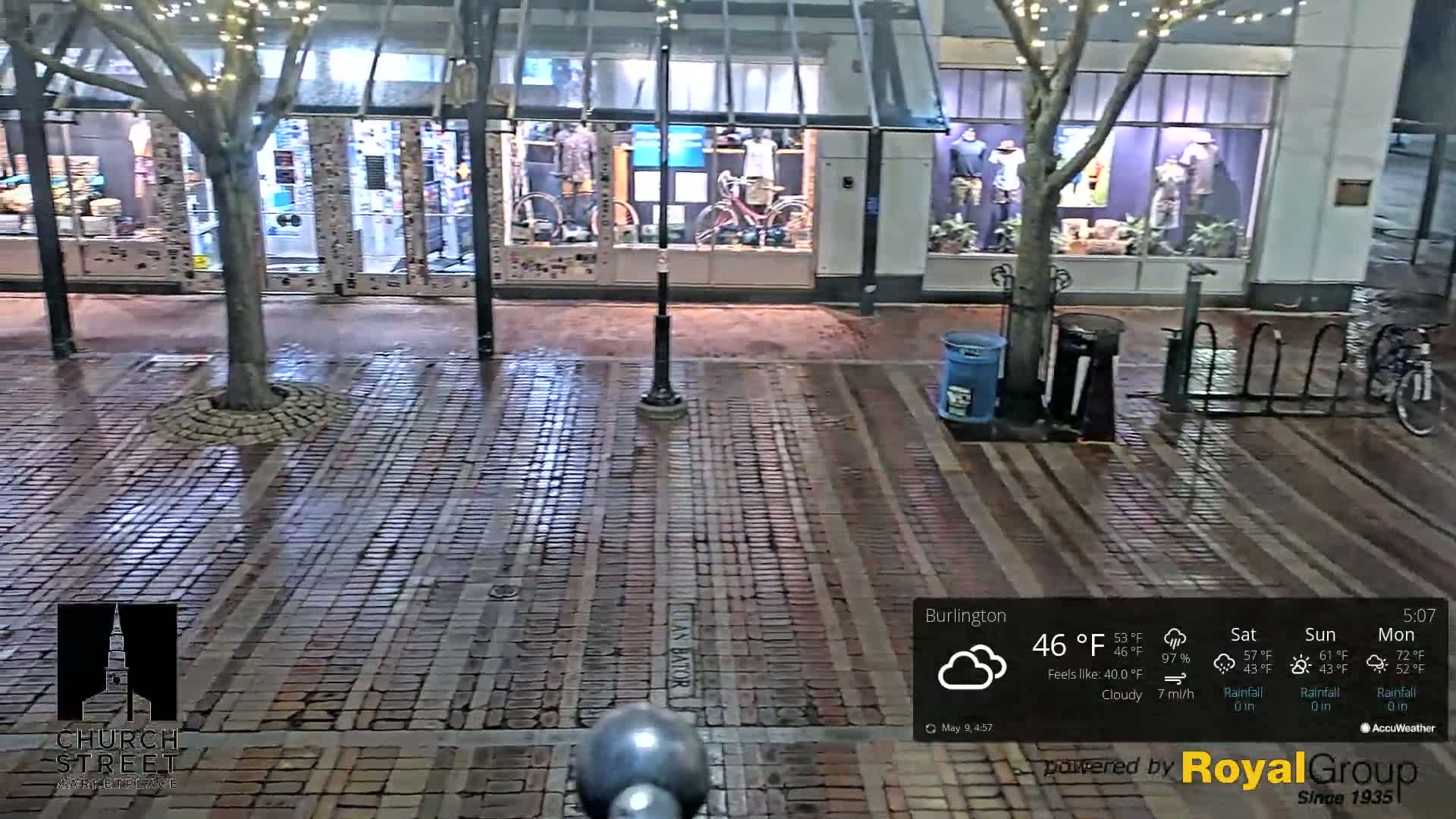 A wet brick street at night, under a cloudy sky, shows store fronts with bicycles displayed in the windows and a bike rack with a bicycle.