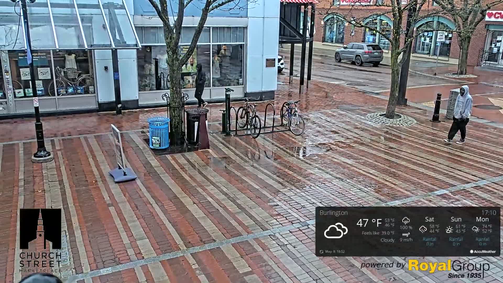 A wet brick street is shown with several pedestrians, bicycles leaning against a rack, and a trash can, under cloudy skies.