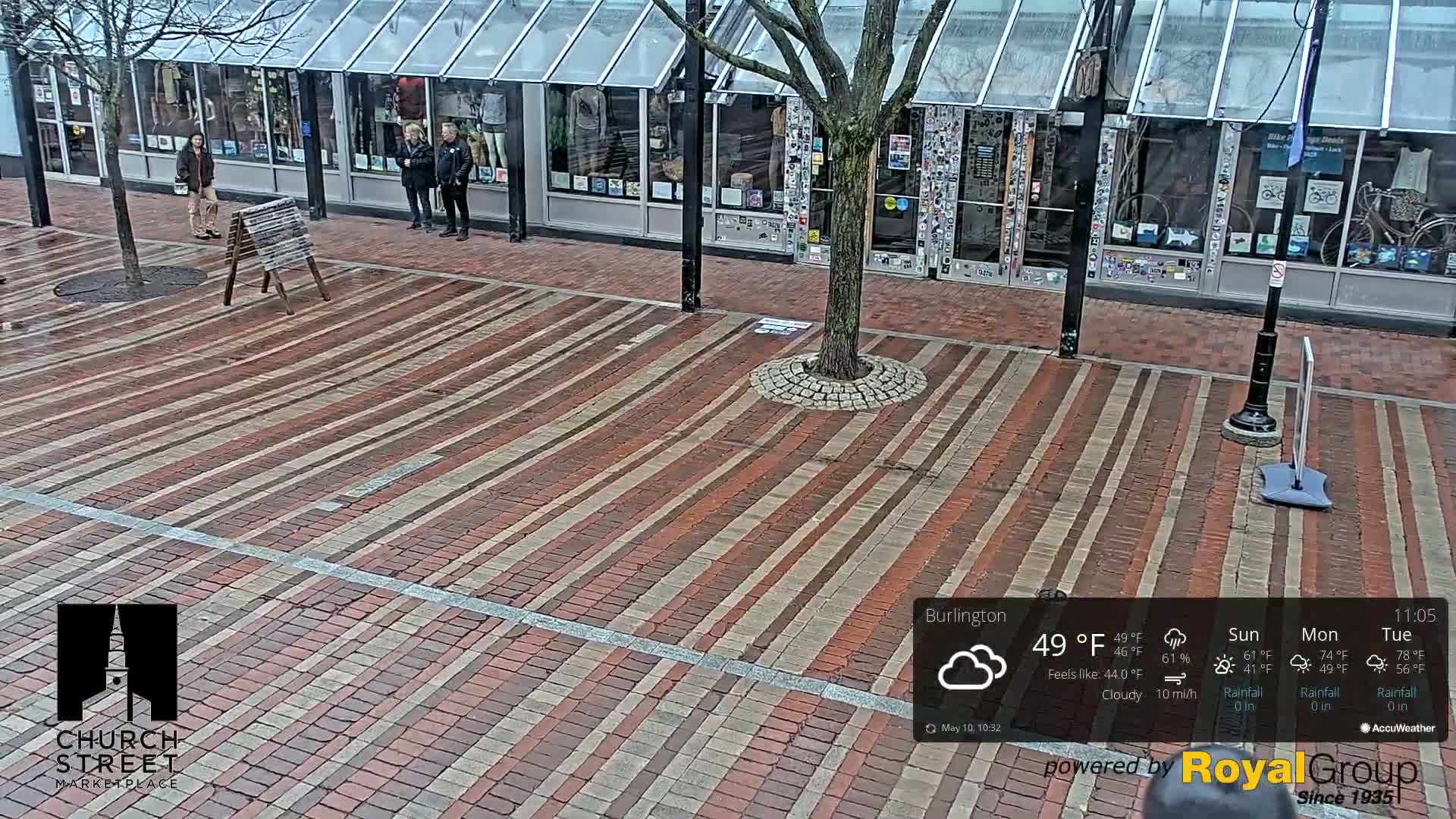A brick plaza with a tree in the center, several people standing near storefronts, and a cloudy day.