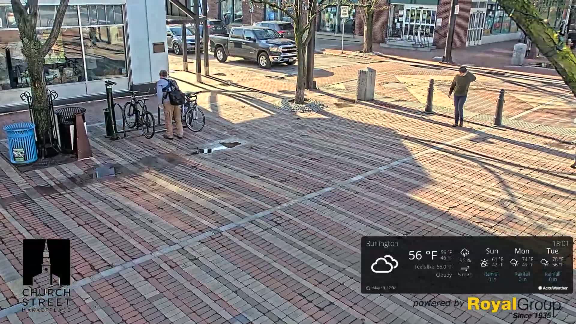 A brick paved area shows two people, one near parked bicycles and another walking further away, under a cloudy sky.