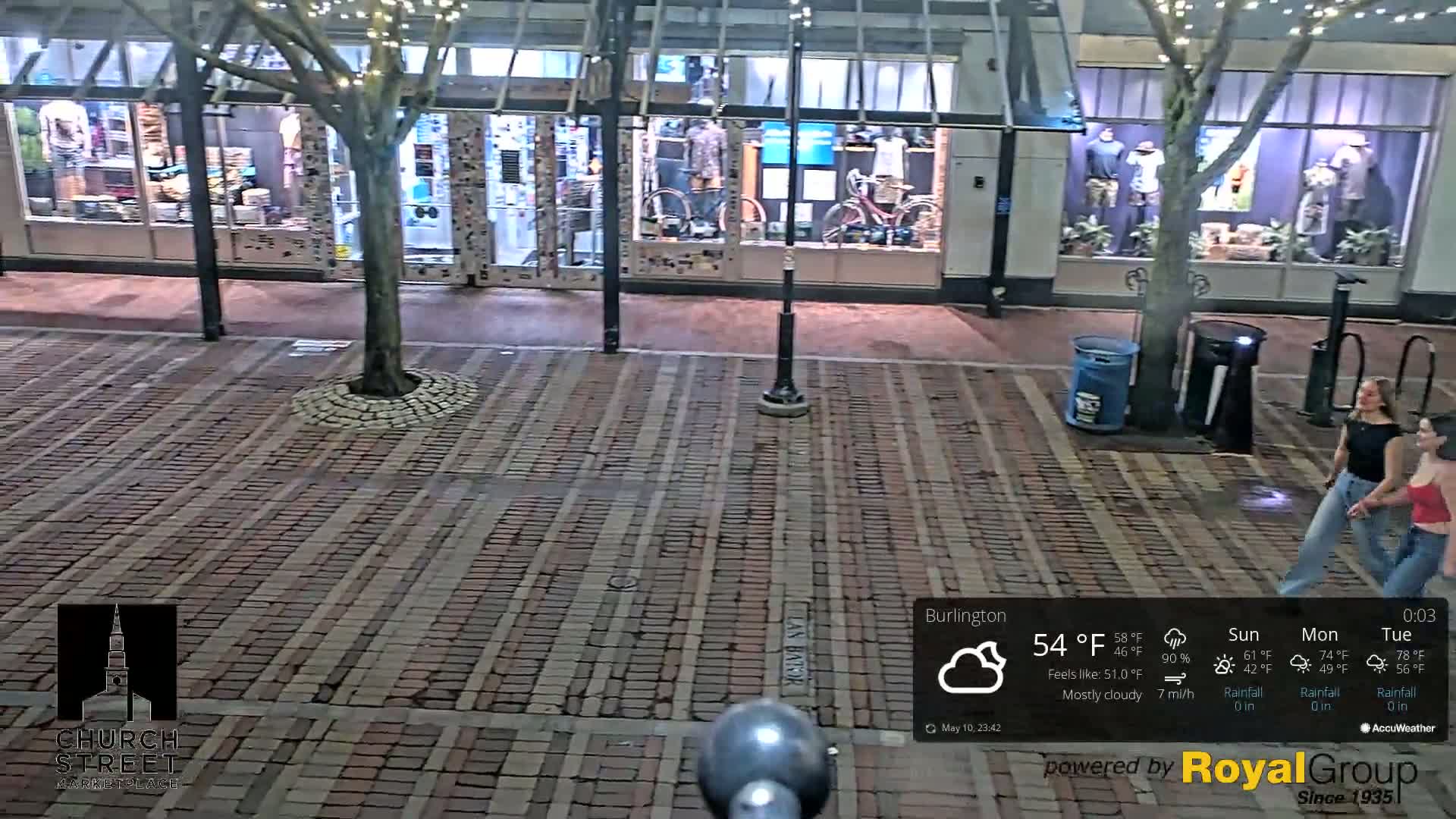 A brick-paved street at night, under mostly cloudy skies, with two women walking past storefronts displaying mannequins and bicycles.