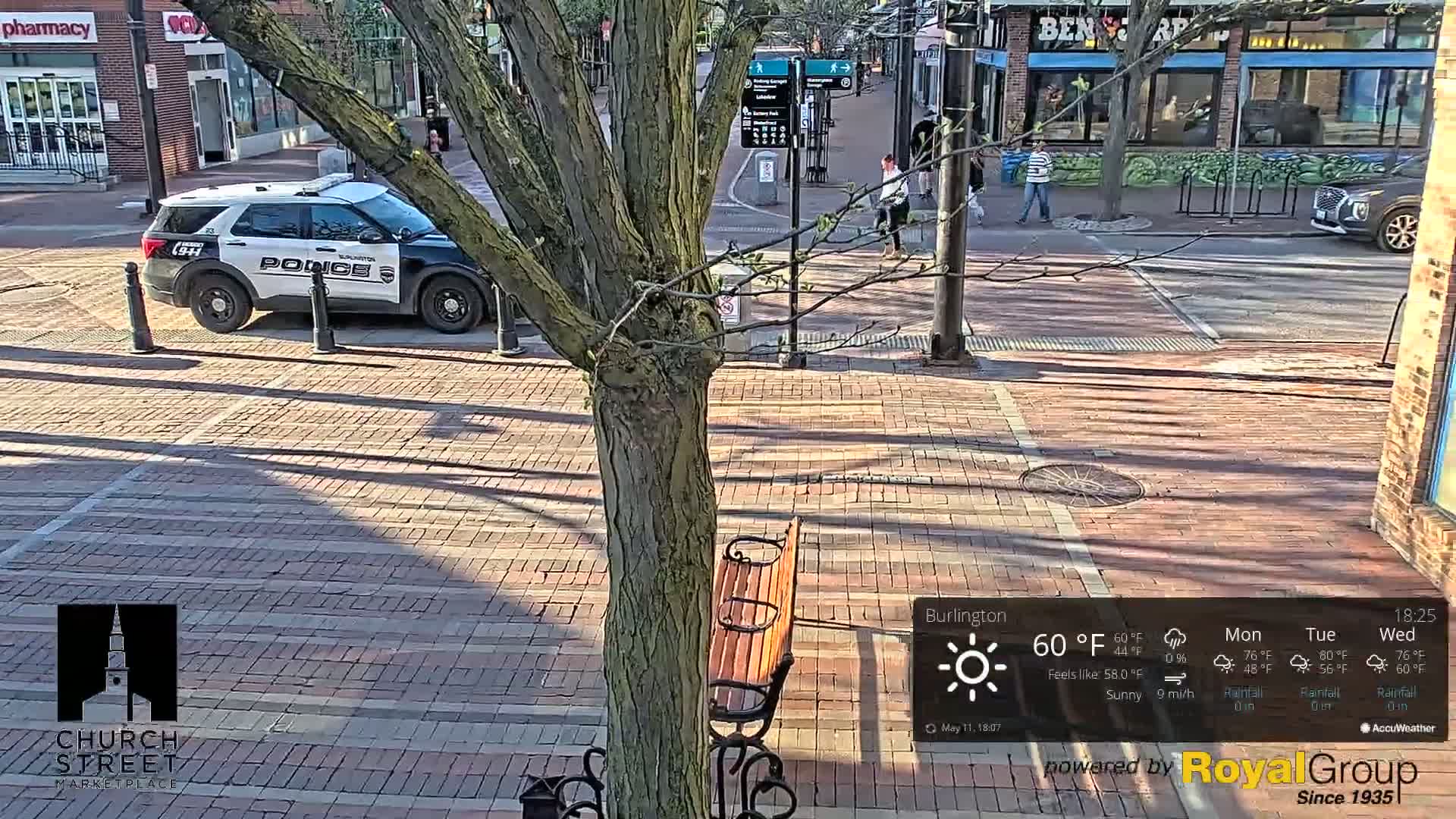 A sunny street scene shows a police car parked near a tree and a park bench, with pedestrians walking on a brick-paved area.