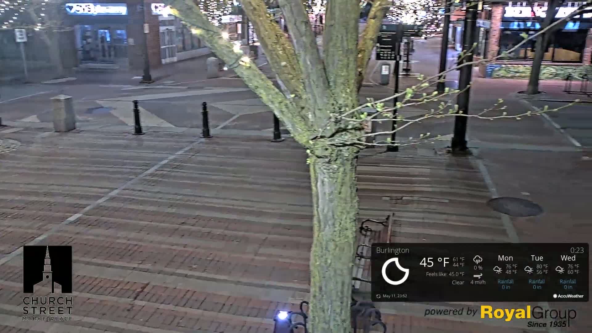 A nighttime view of a brick-paved street with a tree in the center, under clear skies and a temperature of 45°F.