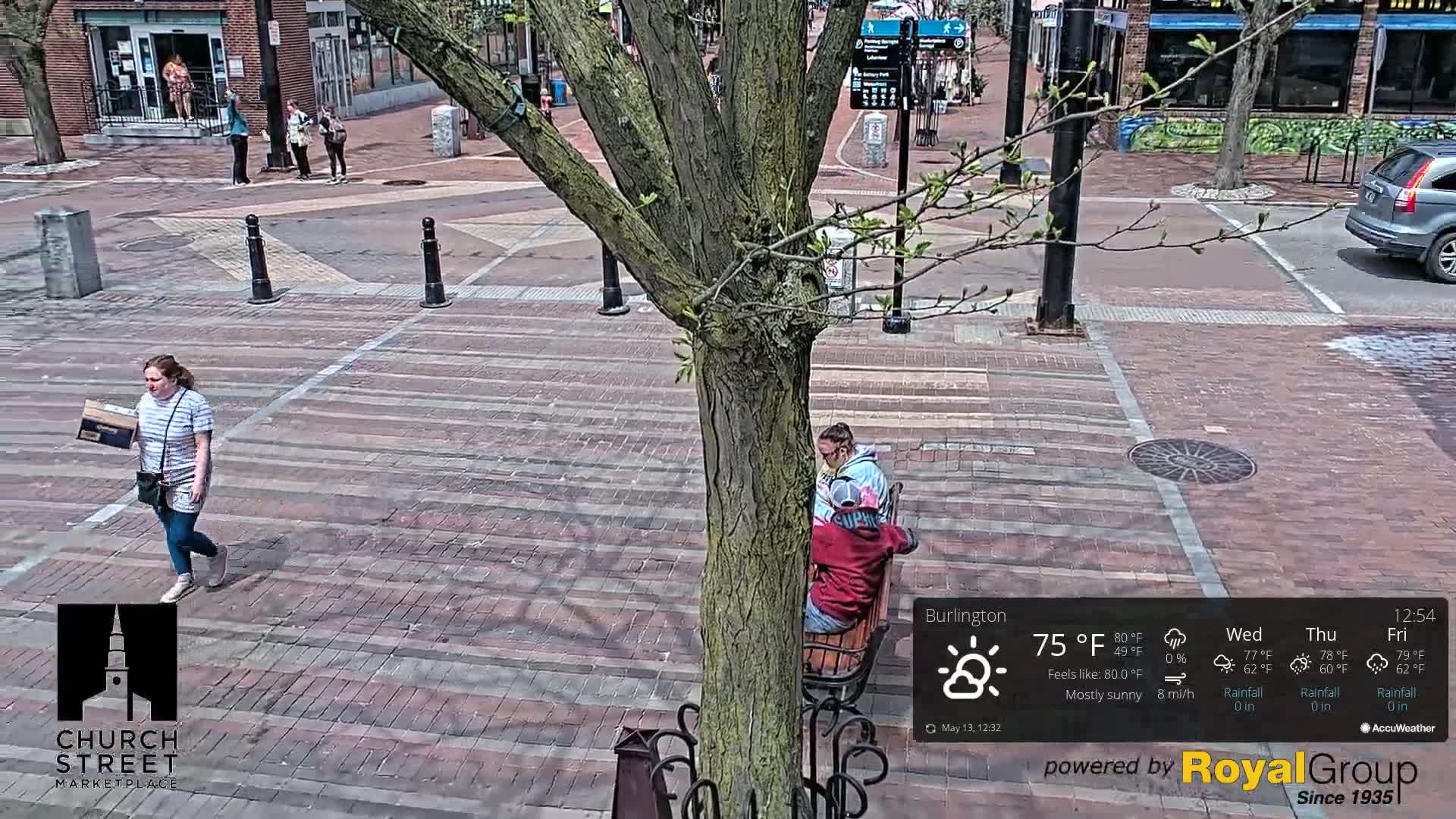 A mostly sunny brick plaza is shown with a few pedestrians and people sitting on a bench near a tree.