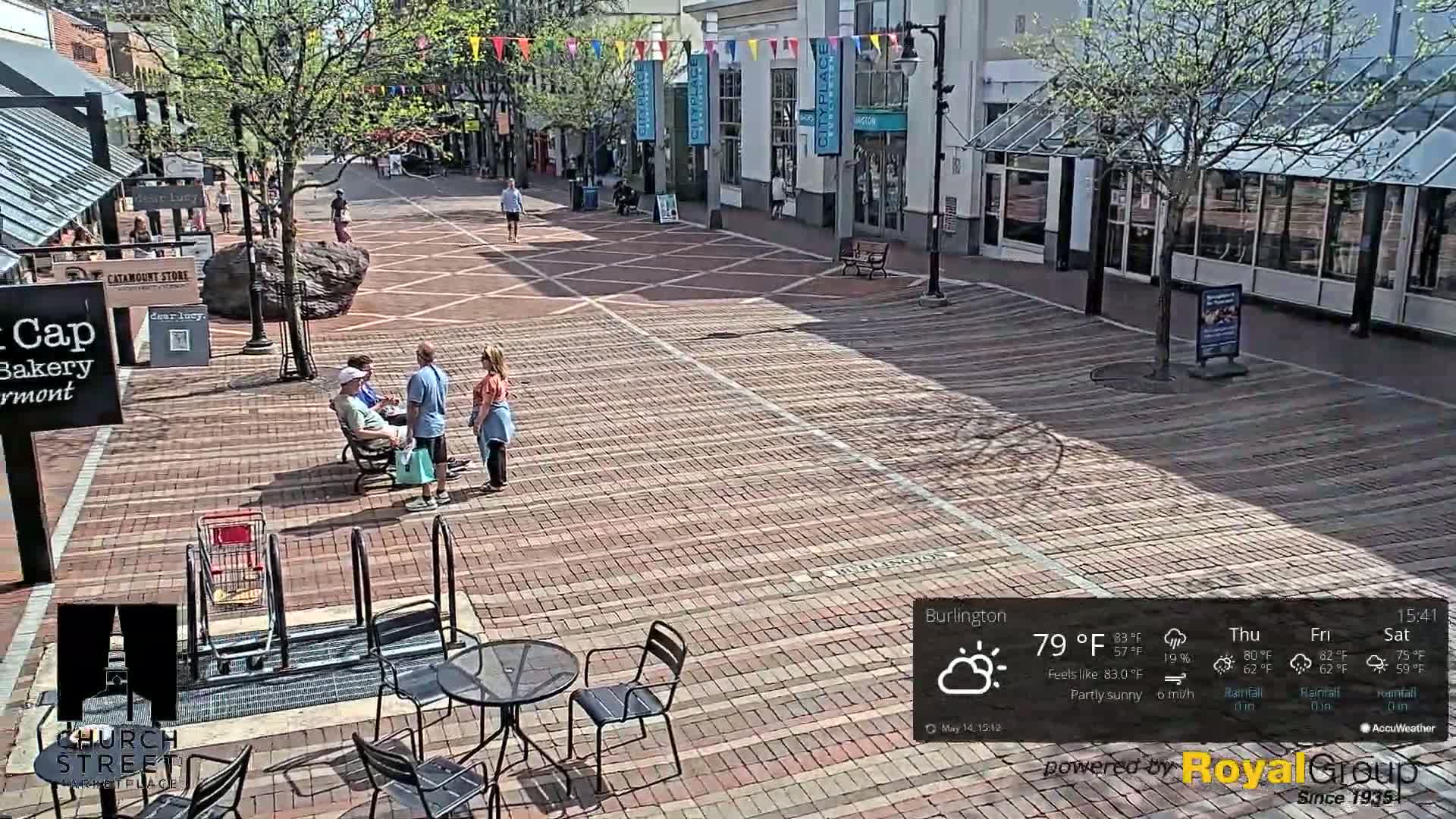 A brick pedestrian mall is partly sunny with a few people walking and sitting, some shops are visible along the sides.