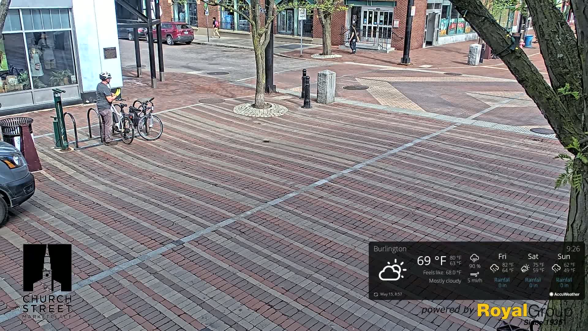 A man stands with two bicycles at a bike rack on a brick-paved street under mostly cloudy skies.