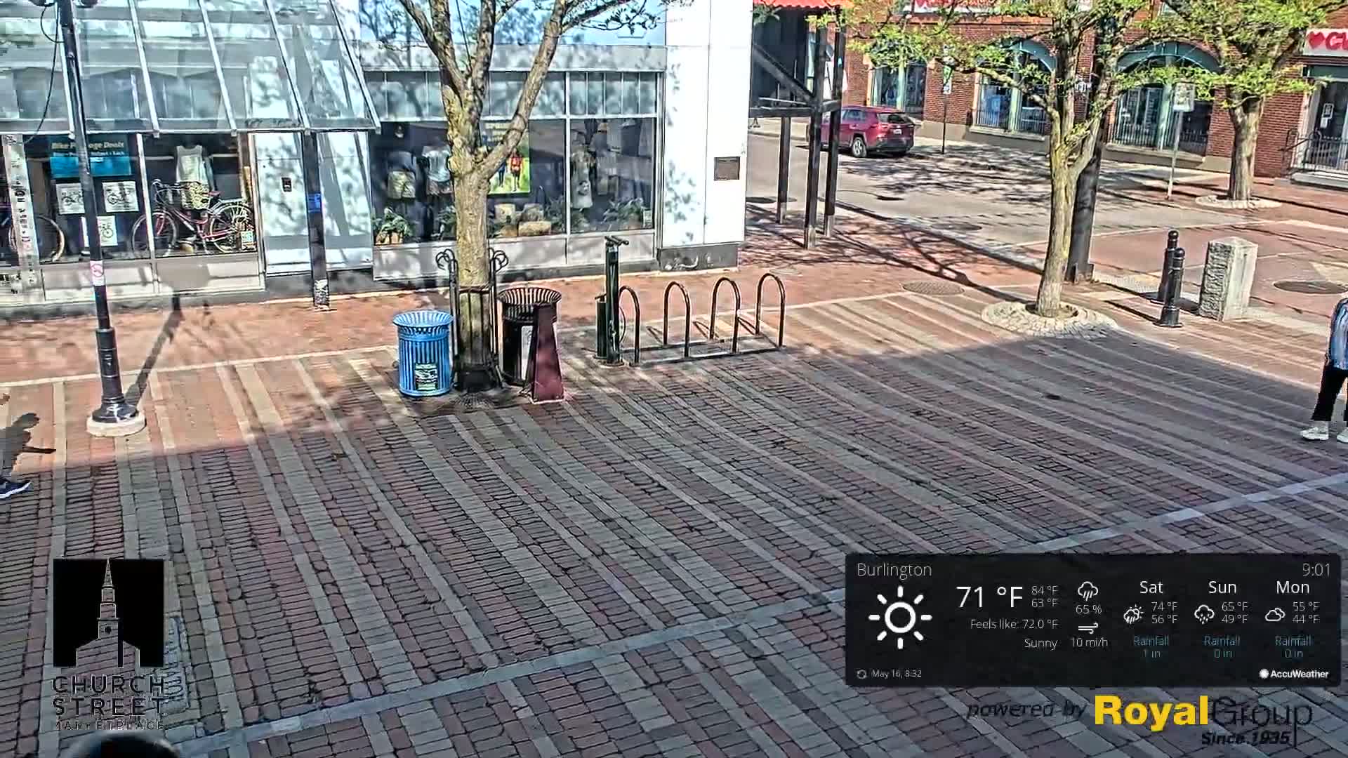 A sunny brick street is shown with a bike rack, trash cans, and a few pedestrians visible.