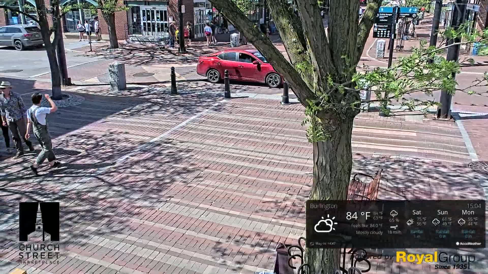 A brick-paved pedestrian area with people walking, a red car, and a tree casting shadows in mostly cloudy weather.